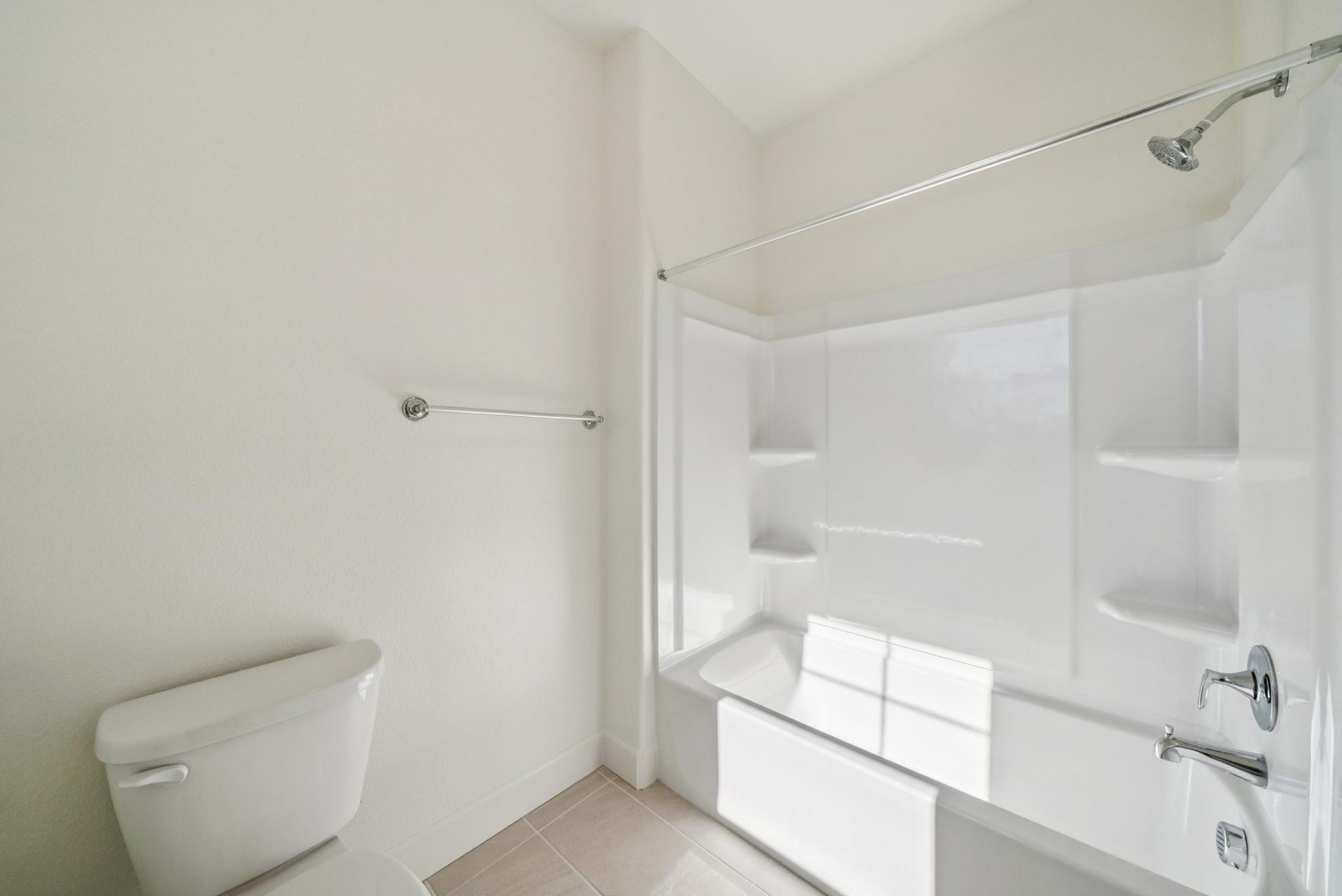 A bathroom with tile flooring with a white bathtub and shower surround with chrome fixtures, and light colored walls.
