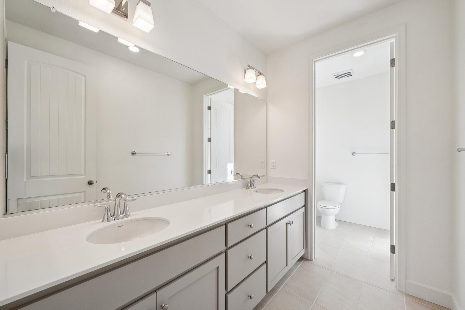 A bathroom long vanity with two sinks, taupe painted cabinets and drawers and a light color countertop with chrome faucets, a large mirror and two light fixtures.
