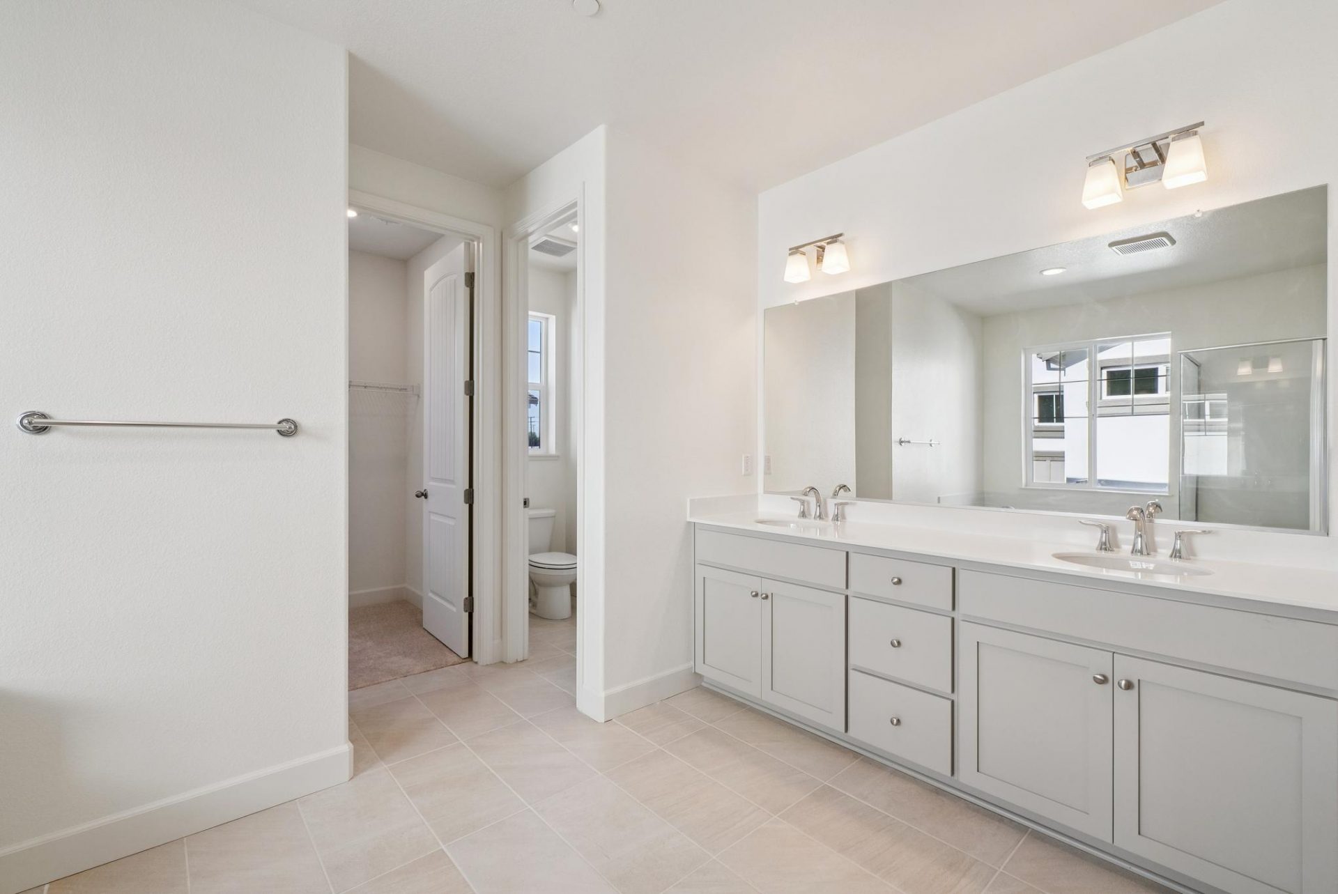 A bathroom with tan tile flooring, a dual sink vanity with a light brown painted base with off-white countertop and chrome faucets with two light fixtures.
