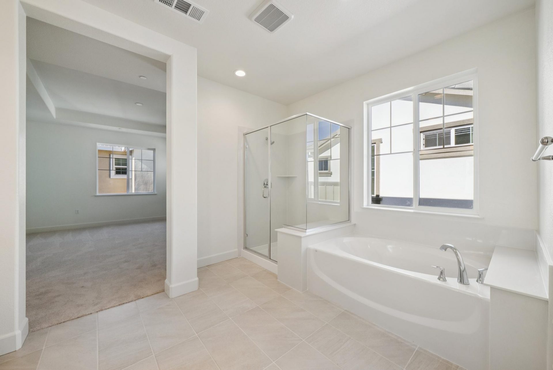 A shower stall with glass enclosure, a soaking tub each with chrome finishes. Above the tub is a large window, the flooring is a light tan tile.