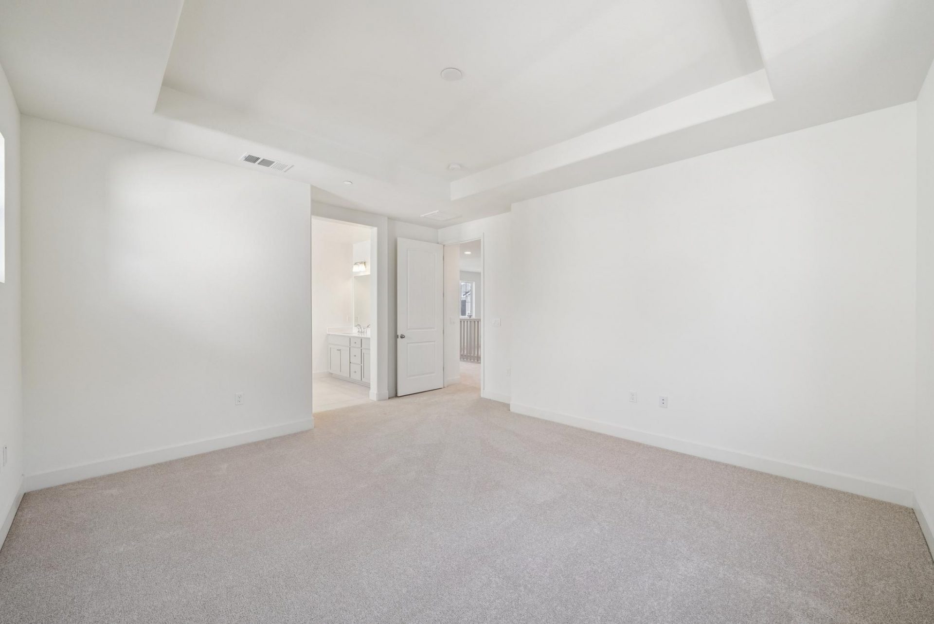 A large bedroom with light colored carpet, and light colored walls with high ceilings, a doorway and a bathroom can be seen in the distance.