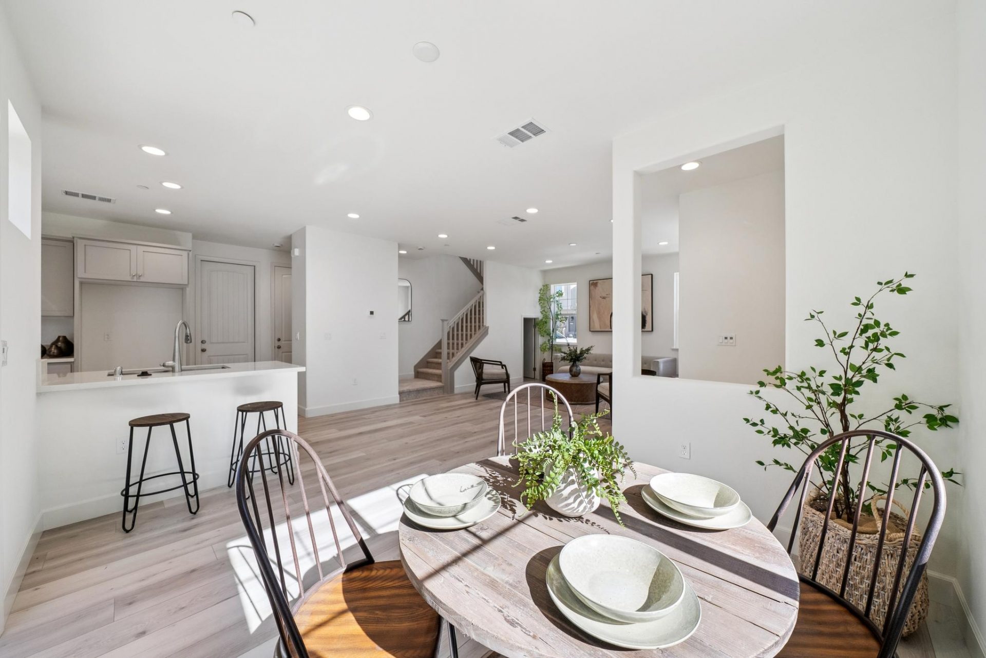 A dining room with a round wooden table and four chairs, table settings and plant décor,  light colored wood tone flooring, and light colored walls. In the distance the kitchen and family room.