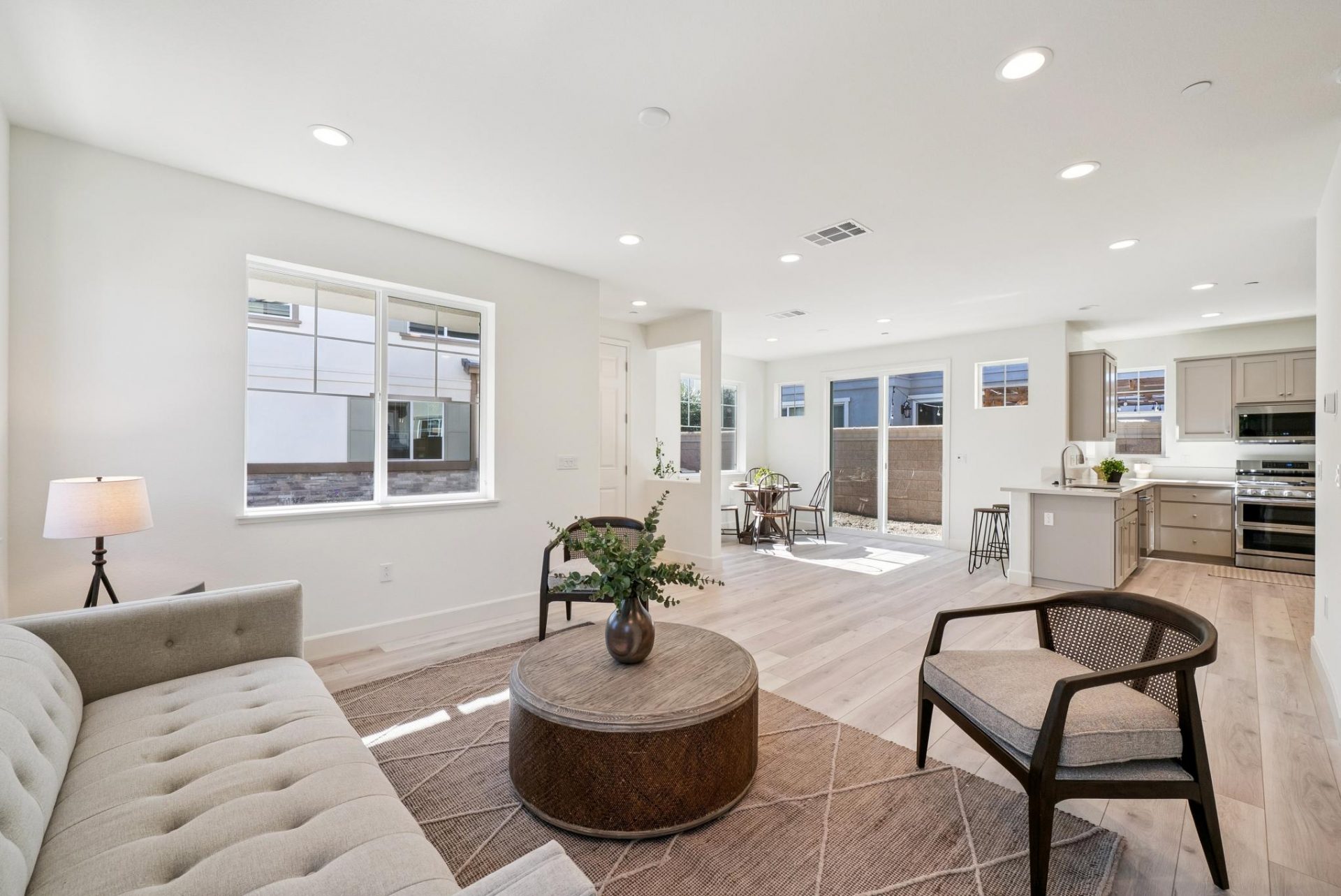 A living area with a couch, chair and a round wooden coffee table, and area rug. Light colored wood flooring seen throughout an open space showing the kitchen and dining area in the distance.