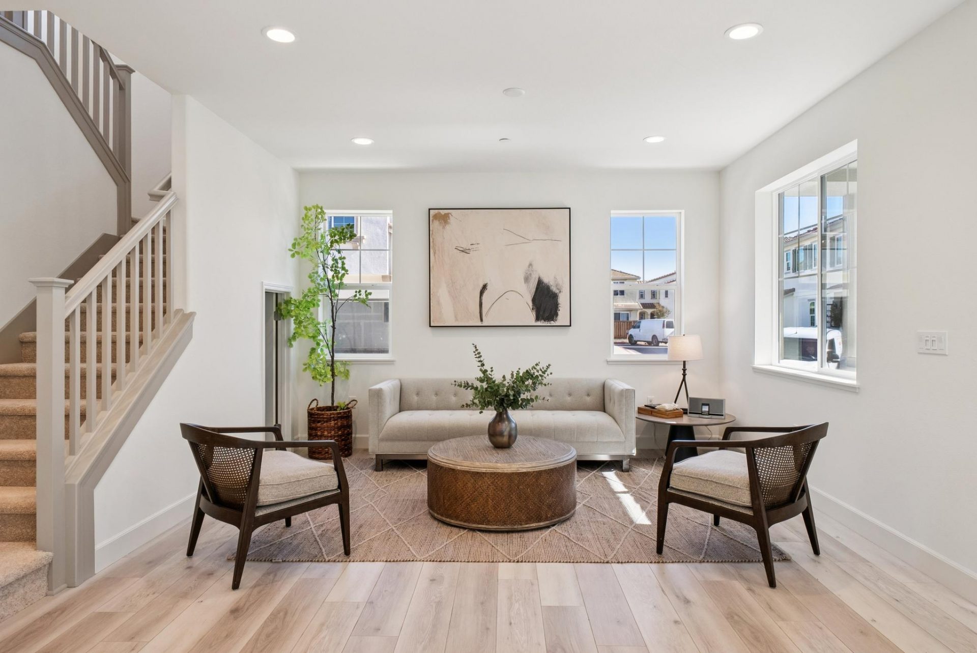 A living area with a couch, chair and a round wooden coffee table, and area rug, light colored wood flooring, plant and wall décor. There are three windows on two adjacent walls.