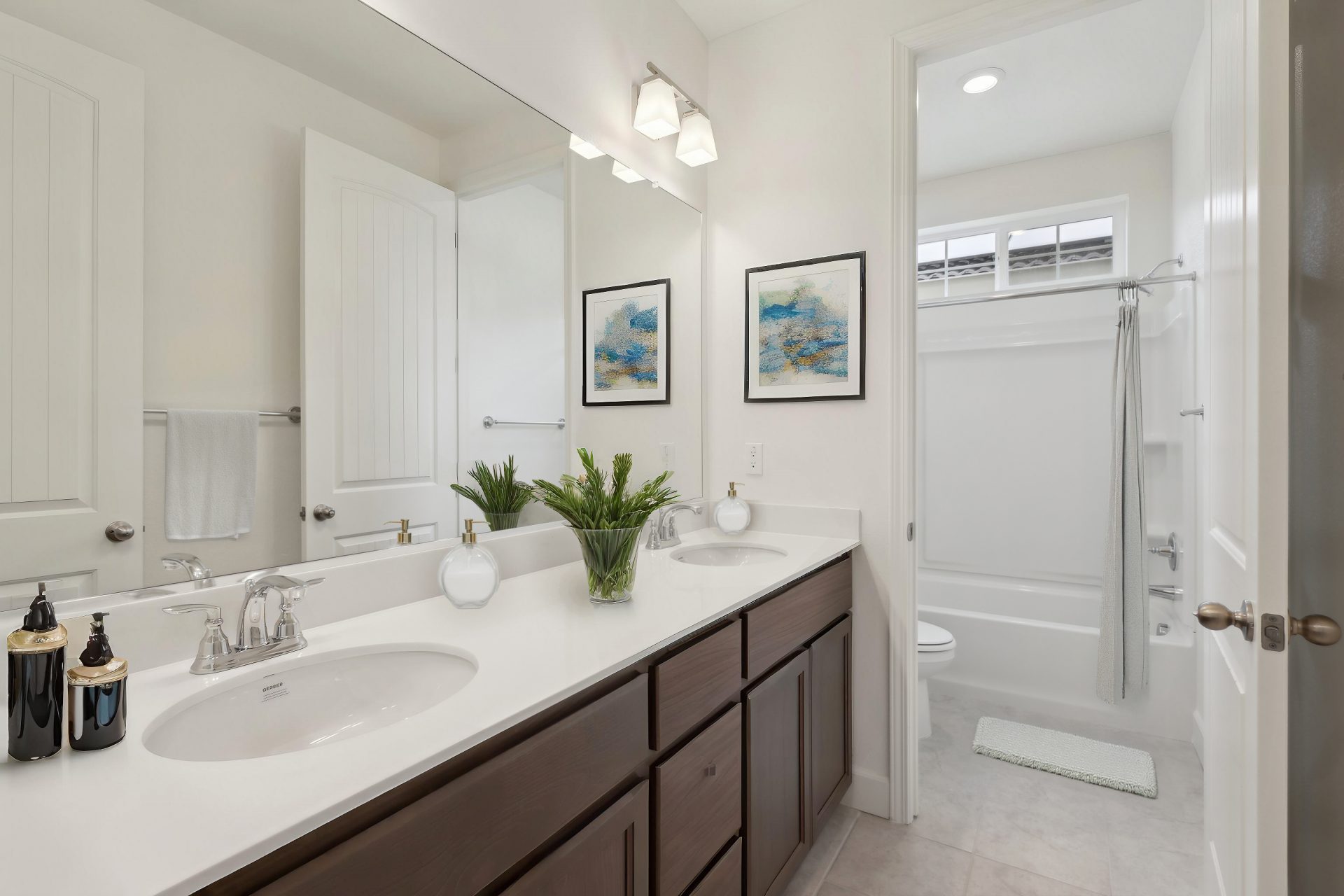 A virtually staged bathroom with light colored tile,  double sink vanity with dark brown cabinets and white countertops, through a doorway a tub shower combo with a toilet.