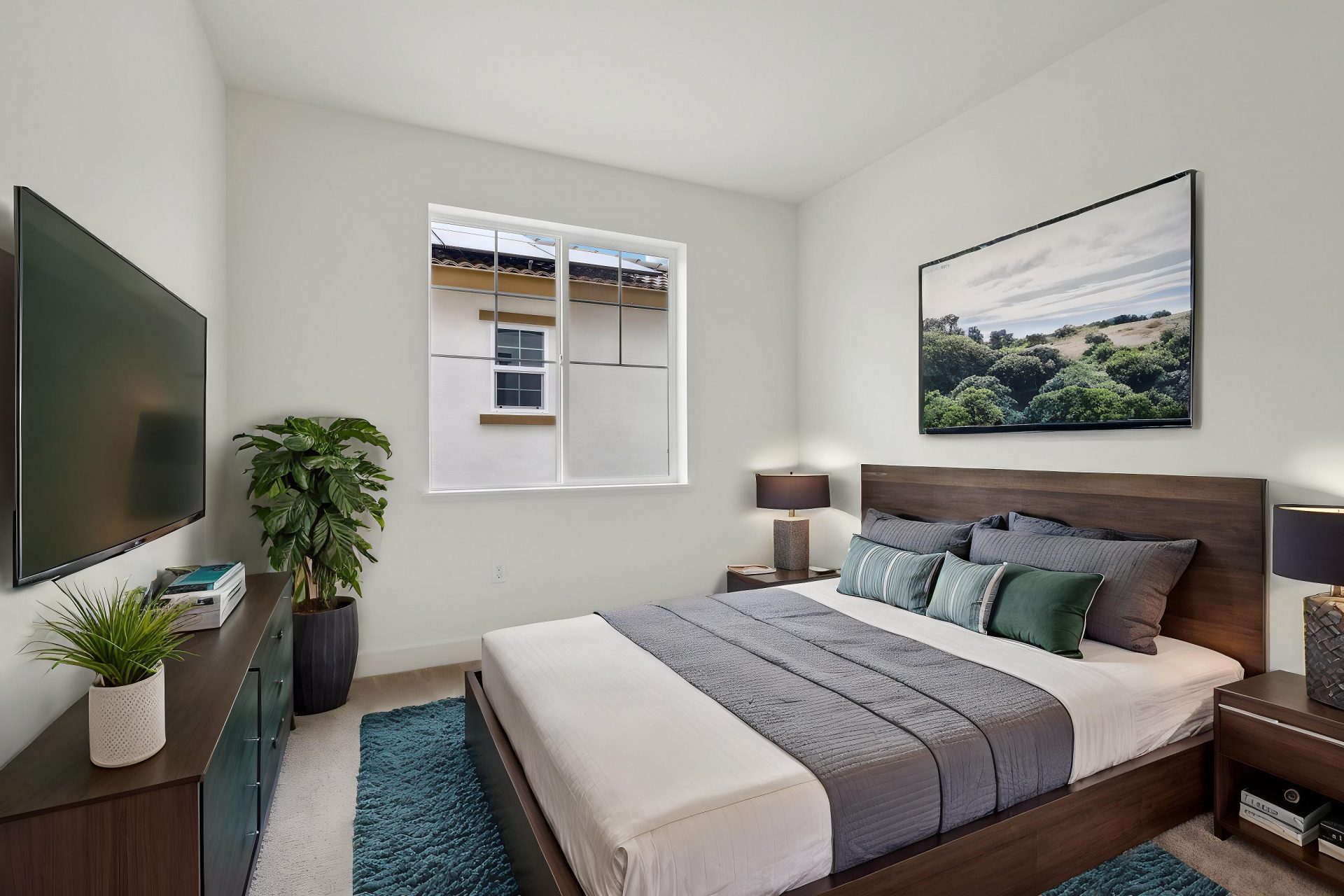 A virtually staged bedroom with light gray carpet, a large window and dark brown furniture, the bed is furnished with blue and tan bedding. To the left is a dresser with a tv above and miscellaneous décor including a potted plant.