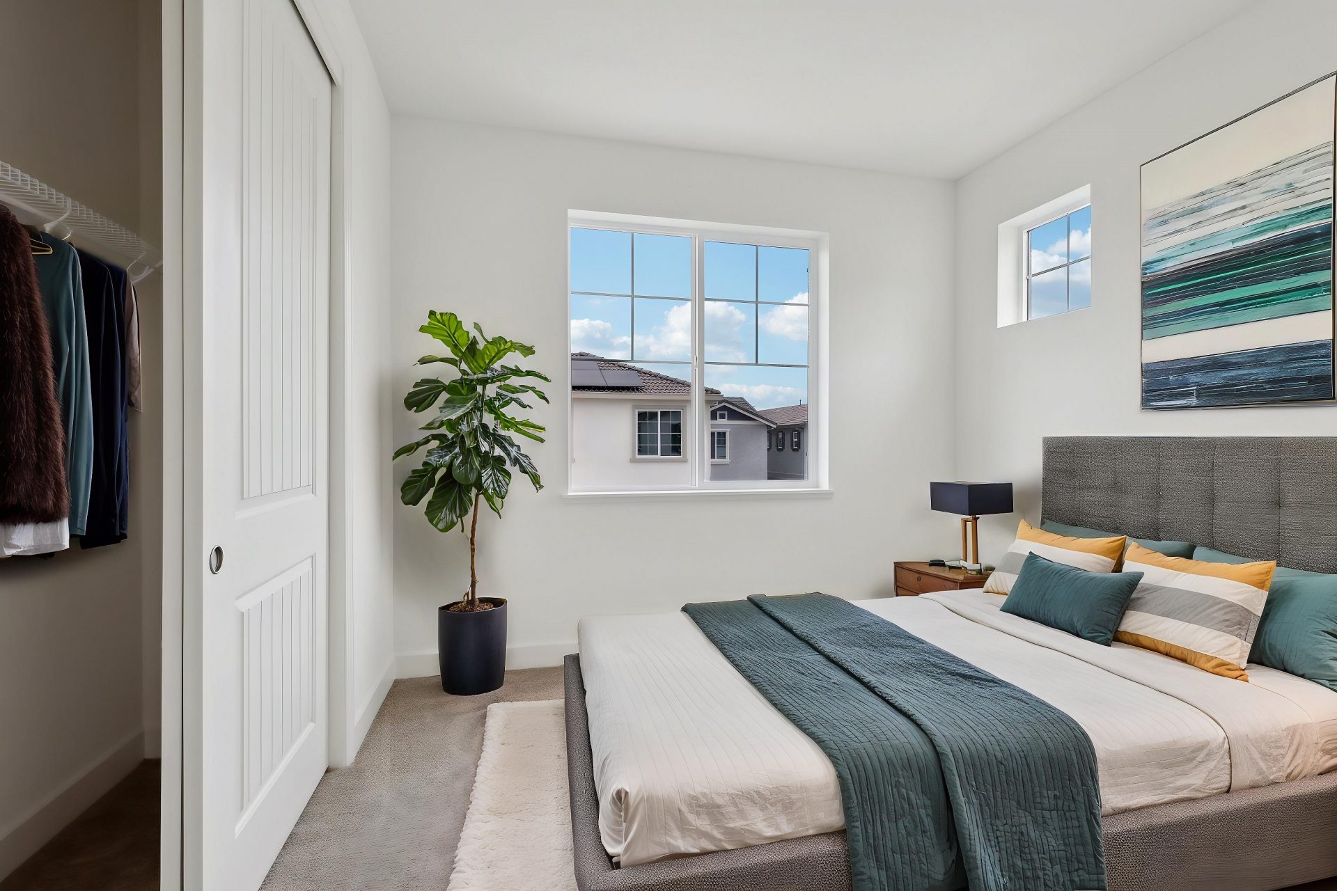 A virtually staged bedroom with a light gray carpet, a bed with gray and turquoise bedding, wall art and a potted plant. As seen are two windows creating a bright space and a closet on the left.