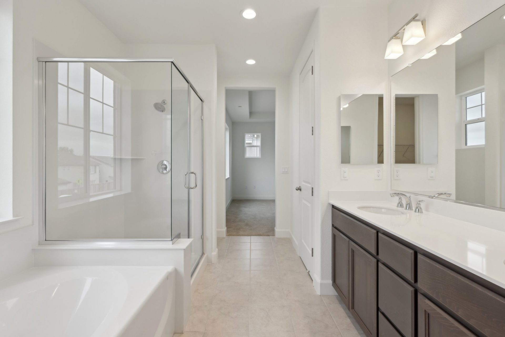 A large bathroom with a soaking tub and separate shower enclosed in glass on the left, and a large double sink vanity on the right with light counters and dark brown cabinets. A large closet can be seen in the distance.