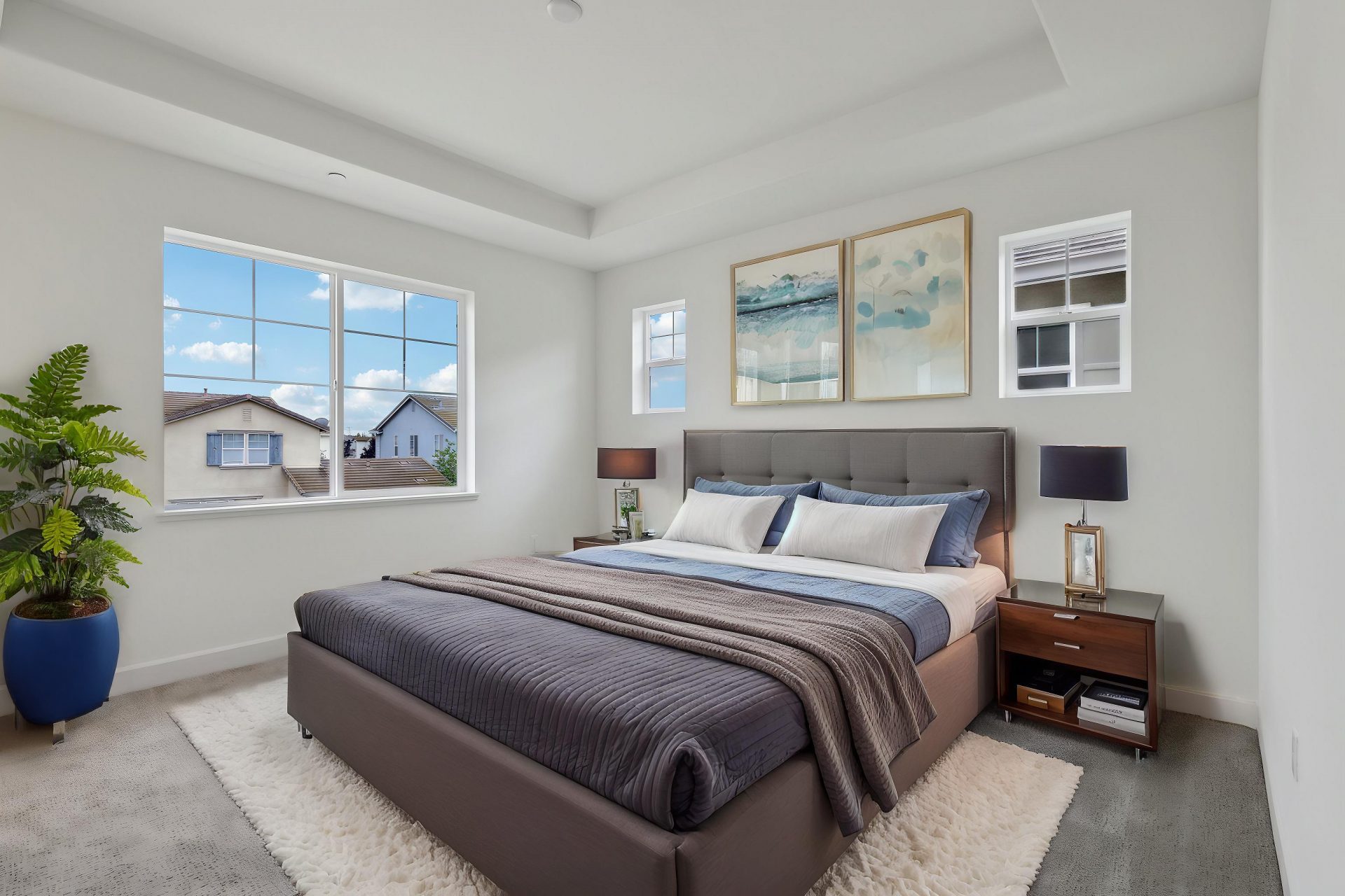 A virtually staged bedroom with gray carpets, staged with a large bed with brown and blue colored bedding, nightstands, lamps, wall art and a potted plant in the corner.