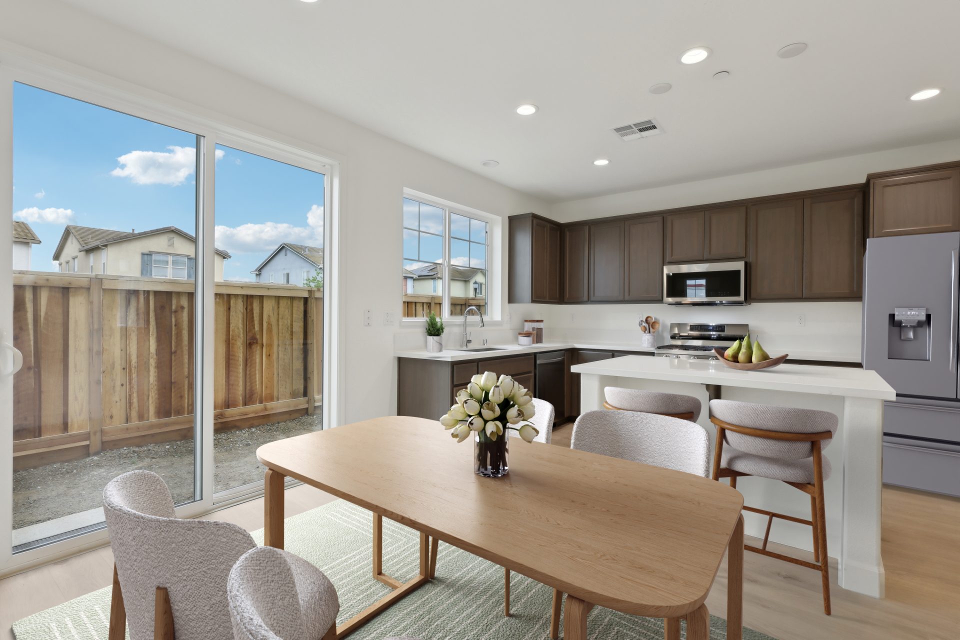 A virtually staged dining area in an open space next to the kitchen and island with white color countertops and darker brown upper and lower cabinets. The flooring is of a lighter wood, and the kitchen is complete with stainless steel appliances.