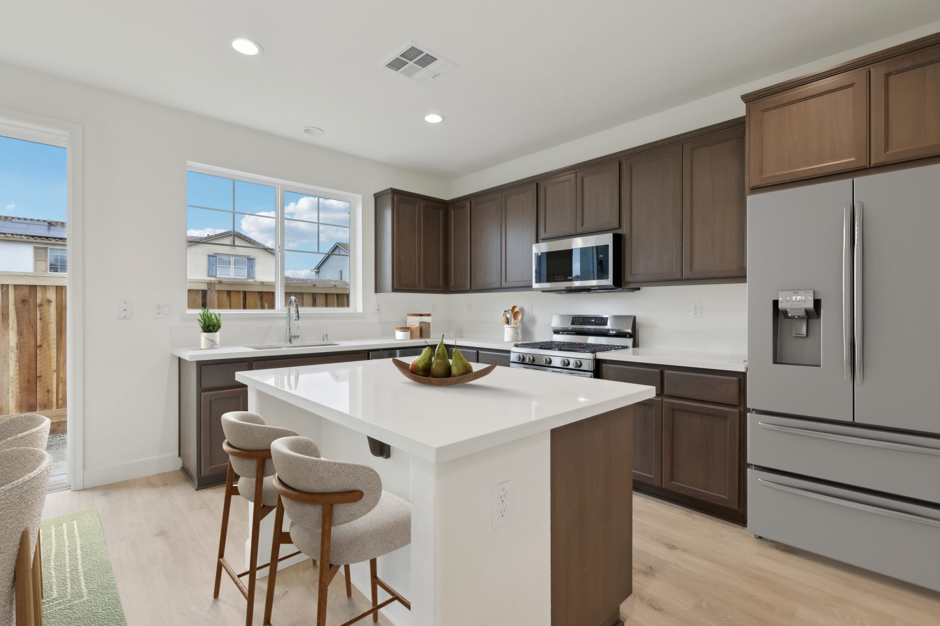 A virtually staged kitchen and island with white color countertops and darker brown upper and lower cabinets. The flooring is of a lighter wood, and the kitchen is complete with stainless steel appliances.