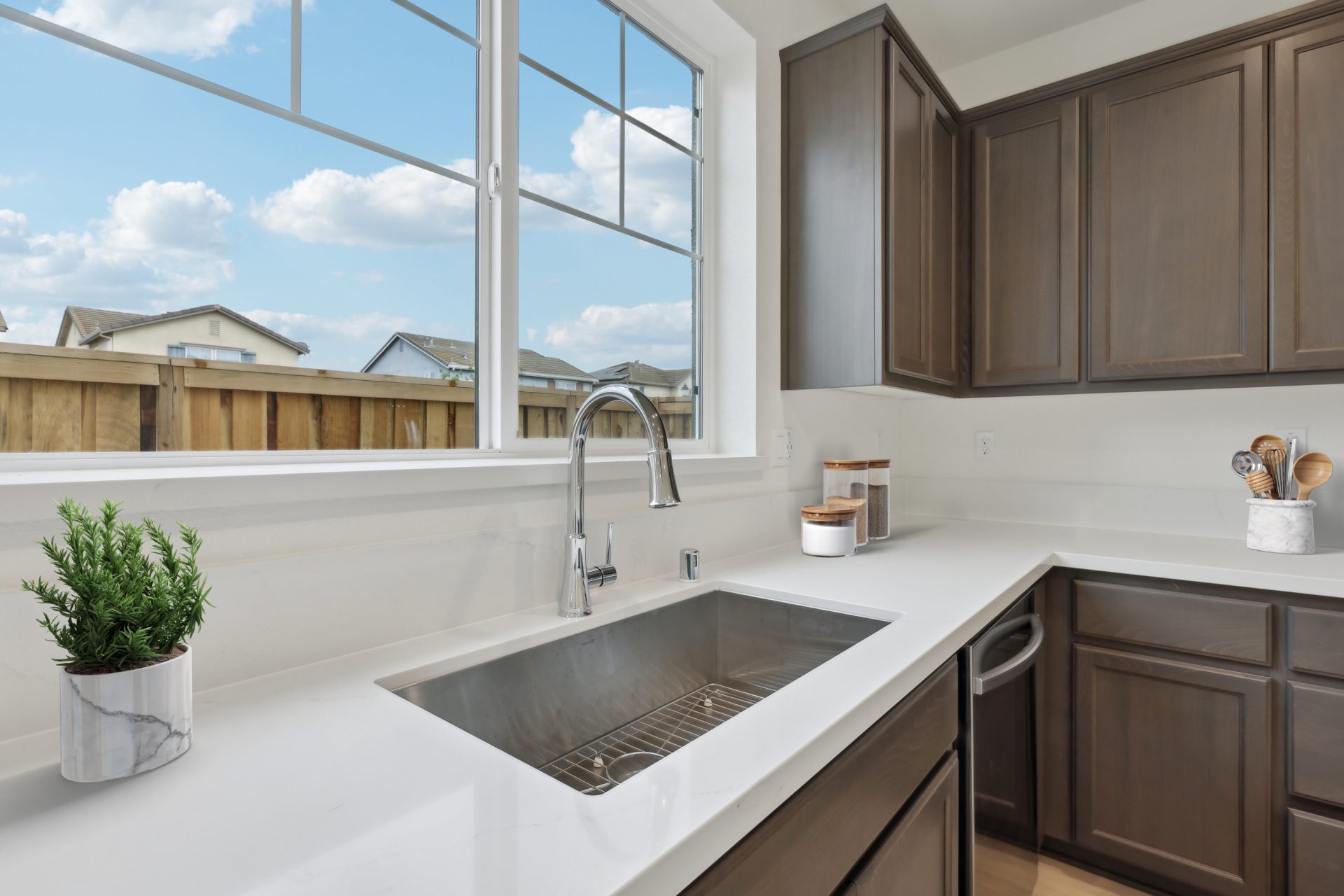 A close up view of the kitchen sink with chrome faucet, virtually staged counter items are seen like a small plant , glass jars, and utensils. There is a large window over the sink .