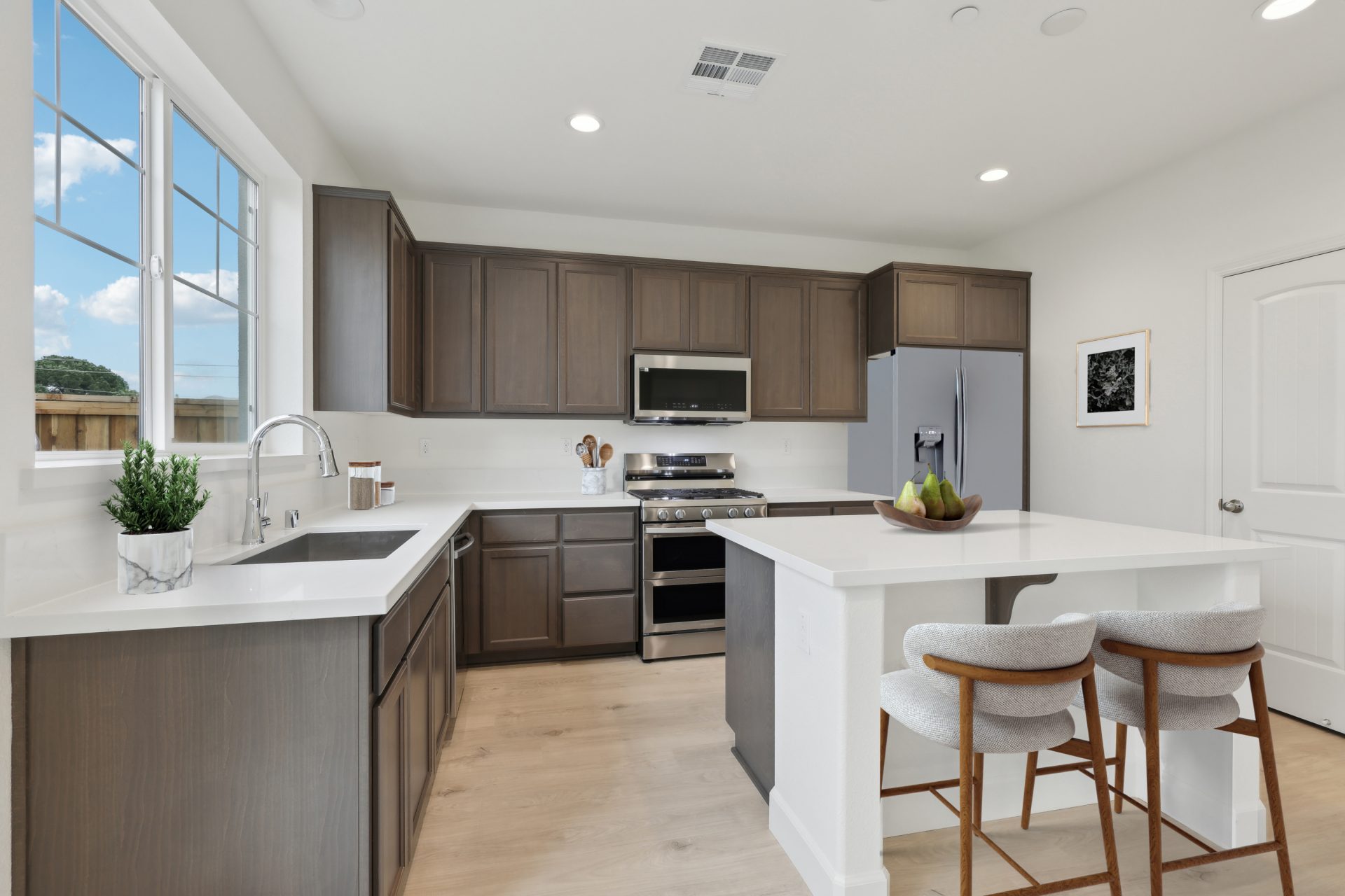 A virtually staged kitchen and island with white color countertops and darker brown upper and lower cabinets. The flooring is of a lighter wood, and the kitchen is complete with stainless steel appliances.