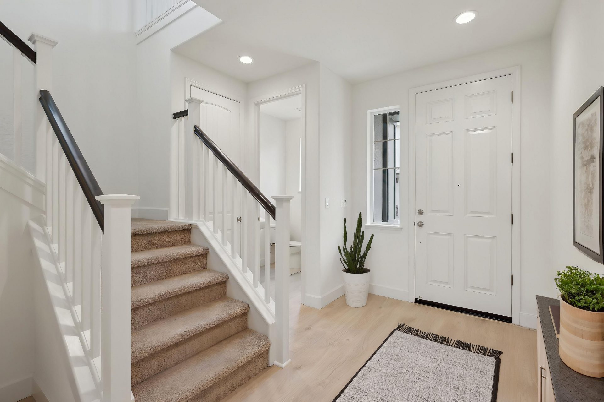 Entry way of a home virtually staged,  with light colored wood flooring, light colored walls and a carpeted staircase with darker stained handrails. In the distance a powder room can be seen.