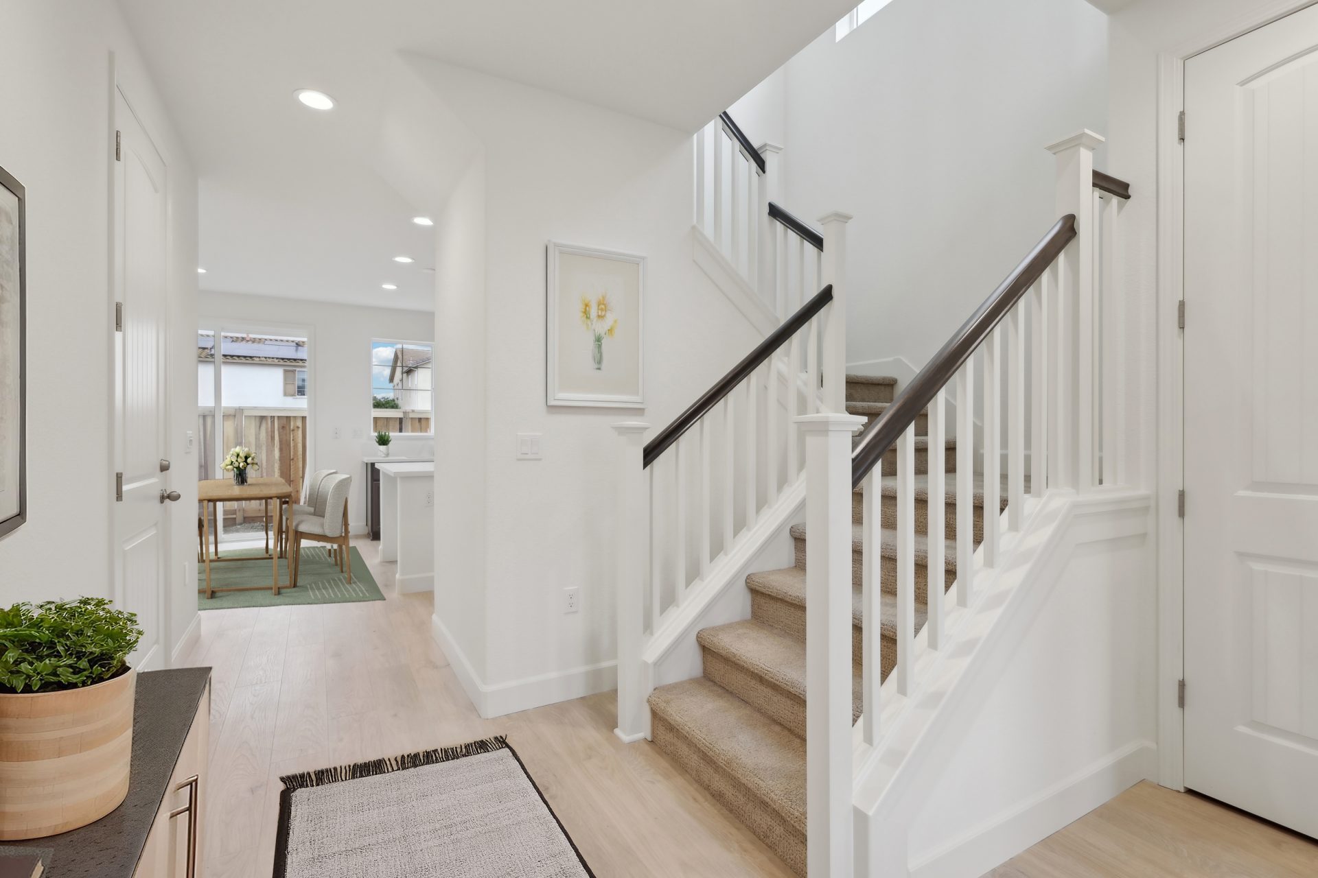 Entry way of a home virtually staged,  with light colored wood flooring, light colored walls and a staircase with darker stained handrails. In the distance a dining area can be seen.