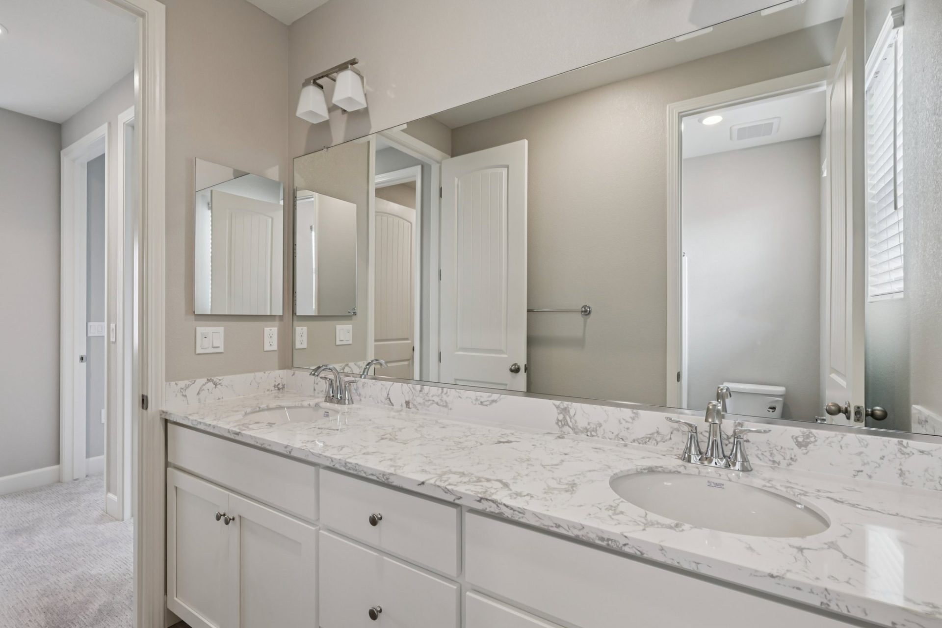 A bathroom with a dual sink vanity a long mirror that spans the length of the vanity and a two-bulb light fixture above the mirror. The sinks have chrome fixtures and there is a mirrored medicine cabinet.