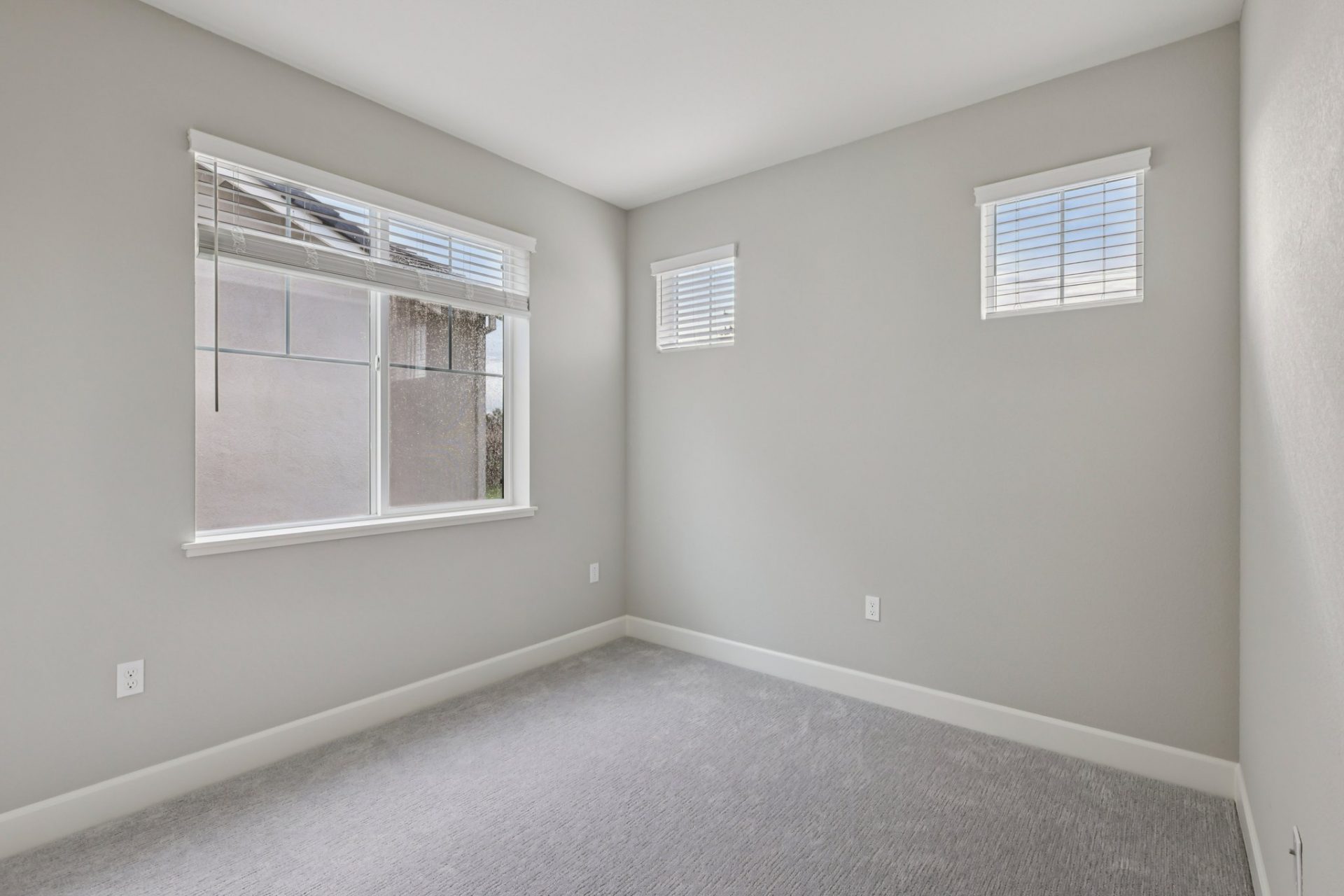 An unfurnished bedroom with a large window, two smaller windows and a closet with two sliding doors, gray carpets and gray colored walls with a white ceiling.