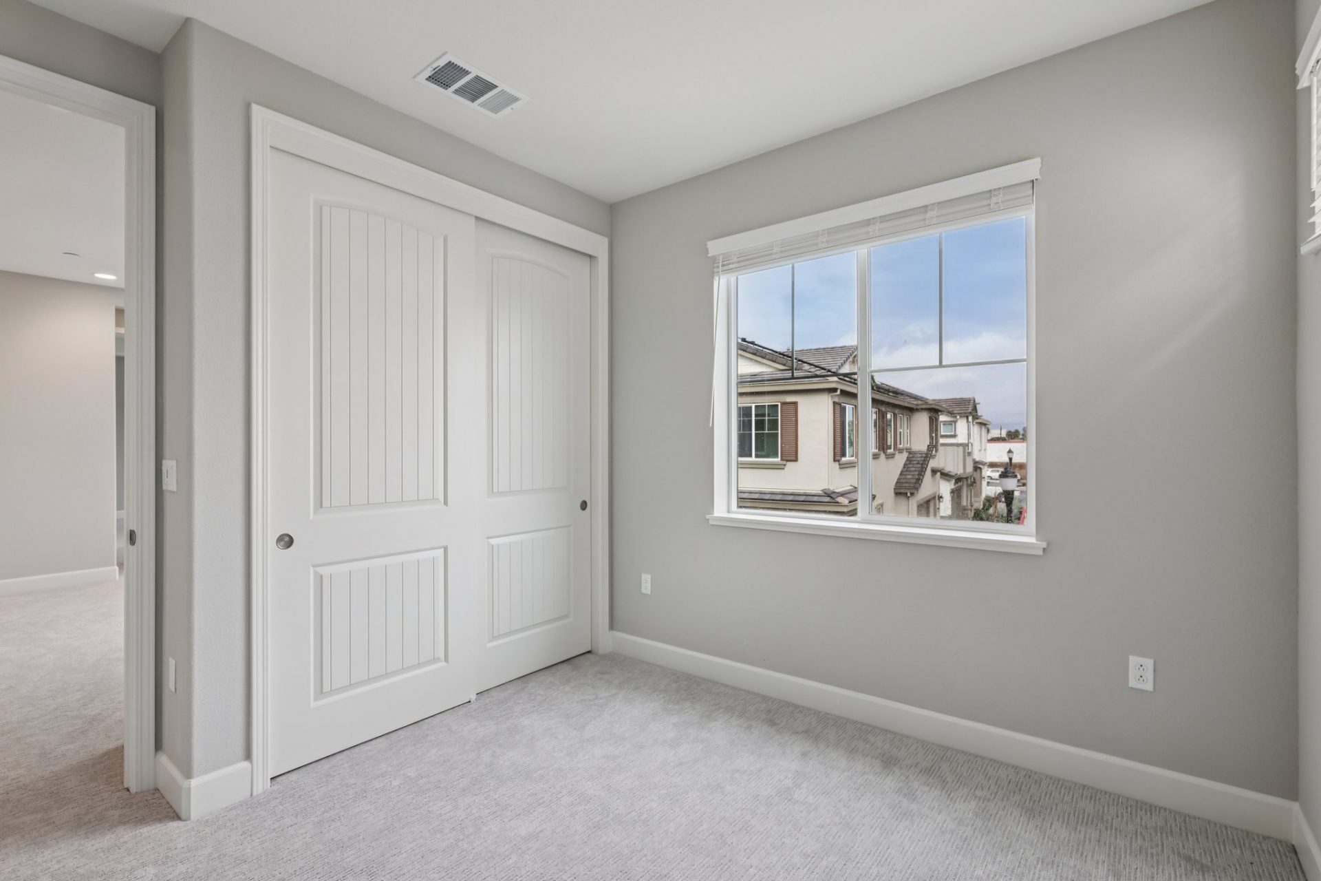 An unfurnished bedroom with a large window and a closet with two sliding doors, gray carpets and gray colored walls with a white ceiling.