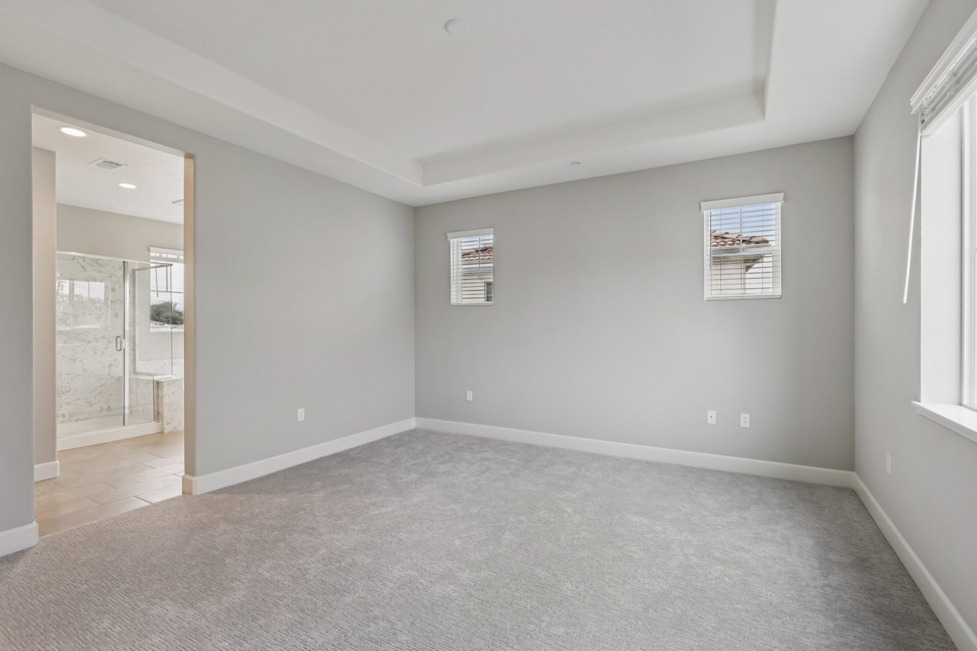 A view of an unfurnished room with two small windows with blinds, to the left an opening that leads to a bathroom.
