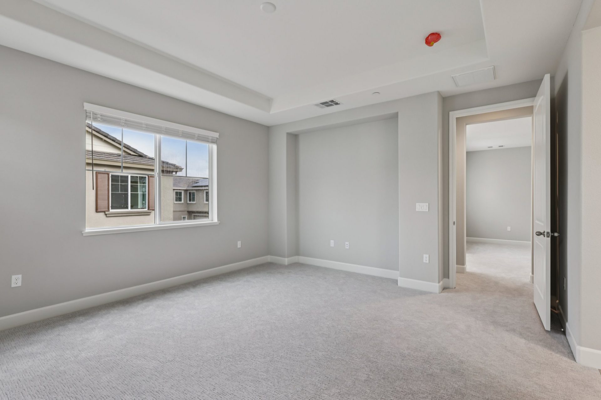 An unfurnished room with gray walls, gray carpets, a large window, and white baseboards. The ceiling and one wall are inset creating some dimension in the room.
