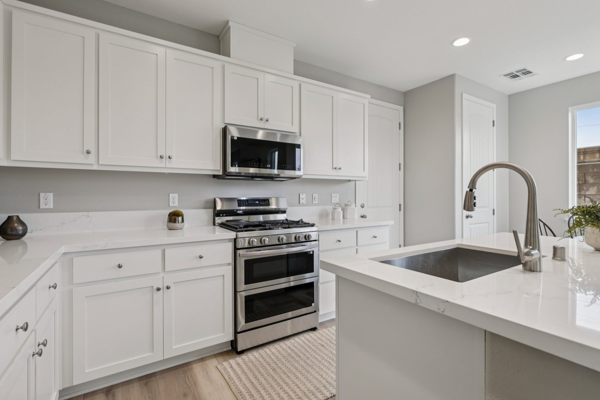 A modern kitchen with white upper and lower cabinetry, quartz countertops, a stainless steel sink in the kitchen island with a nickel color faucet and pull knobs. There is a stainless steel oven with microwave hood setup.