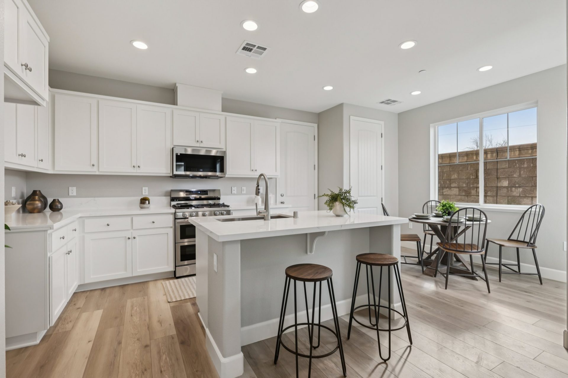 A modern kitchen with an island, white countertops and white cabinets with nickel finishes and stainless steel appliances. Flooring is light toned wood, the rooms are staged with bar stools, and a dining room table with four chairs and various pieces of d
