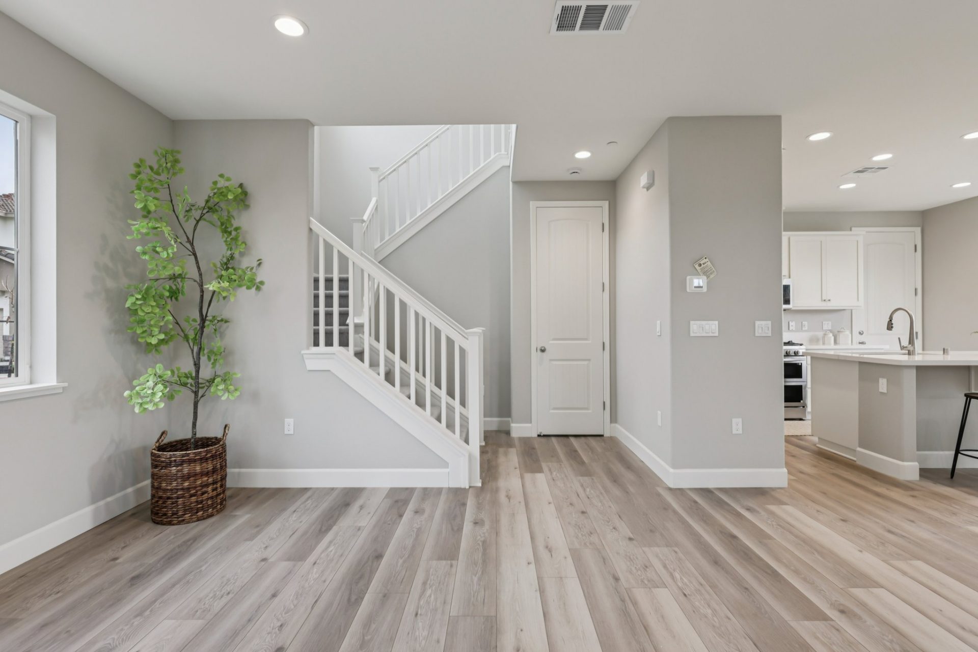 An open space in the living area leading to the stairway with a potted tree on the corner and  light color wood flooring. To the right of the photo the kitchen can be seen in the distance.