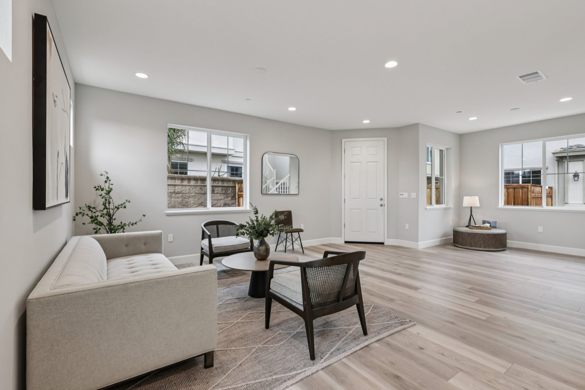 A staged living area with a light color couch, round table and two seating chairs, light wood flooring, medium gray color walls and potted plant décor, two windows on either side of the room and other décor items can be seen.