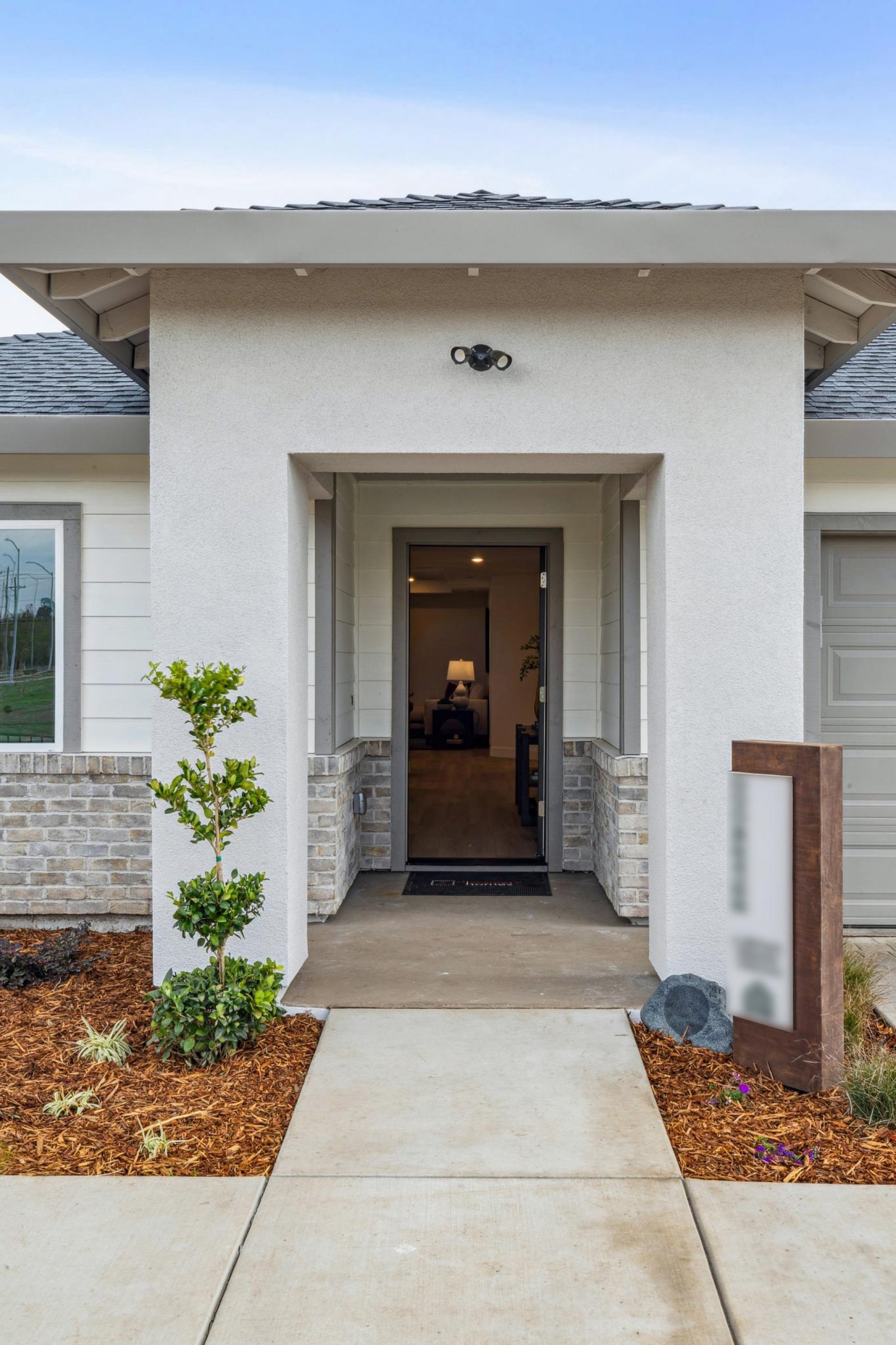 An exterior covered entry way with mulch and shrub landscaping with light gray paint color and stone detail at the front. In the distance an open front door.