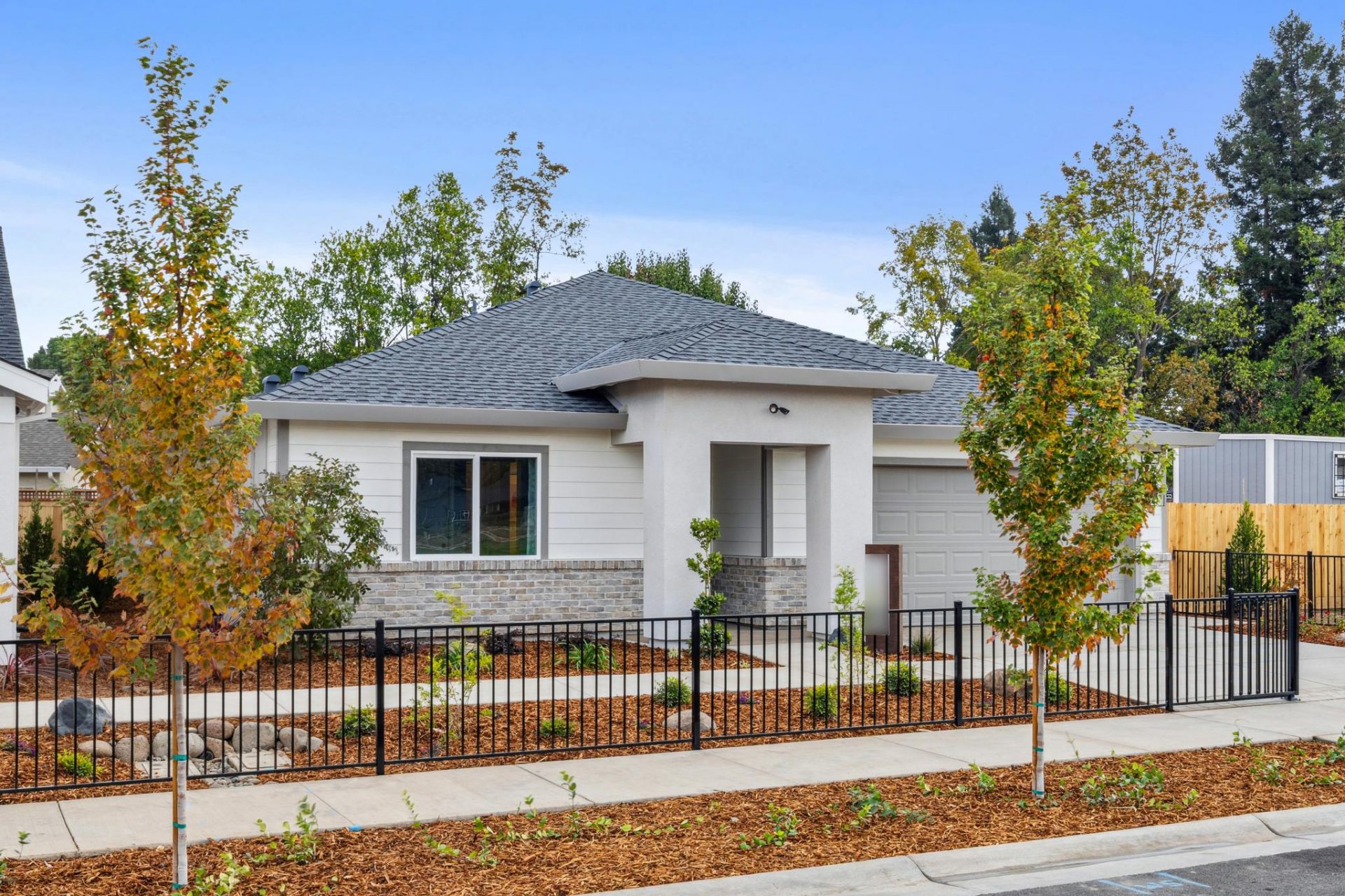 An angled view of the exterior of a single story home with gray paint color, stone detail at gray color roof. Brown mulch landscaping with shrubbery and small trees.