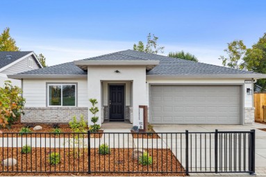 A straight forward view of the exterior of a single story home with gray paint color scheme, stone detail at gray color roof. Brown mulch landscaping with shrubbery and small trees.