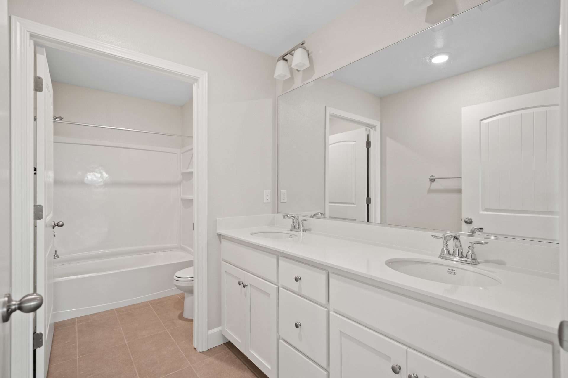 A bathroom with brown tile, white double sink vanity with white countertops and chrome fixtures, long mirror with a tub surround and toilet separated by a wall and door.