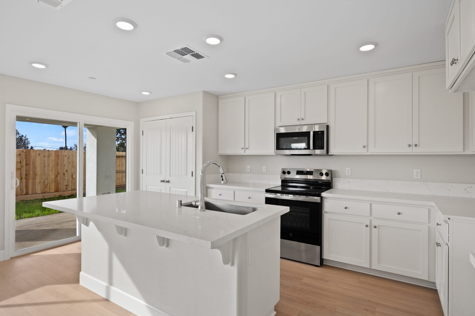 A modern kitchen with light color upper and lower cabinets, light colored countertops with stainless steel appliances, chrome sink faucet and a double door pantry.