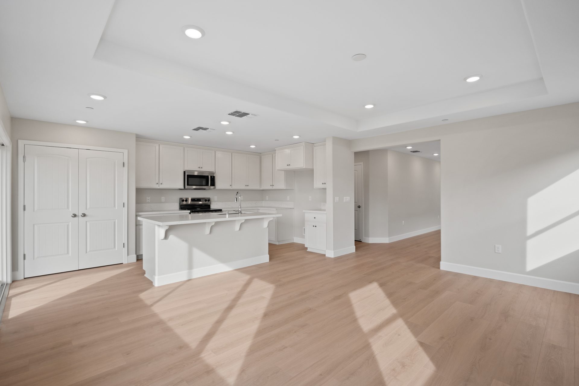 An open concept room space of a family room and kitchen. Light colored wood floors, medium color walls and light cabinets in the kitchen with stainless steel appliances.