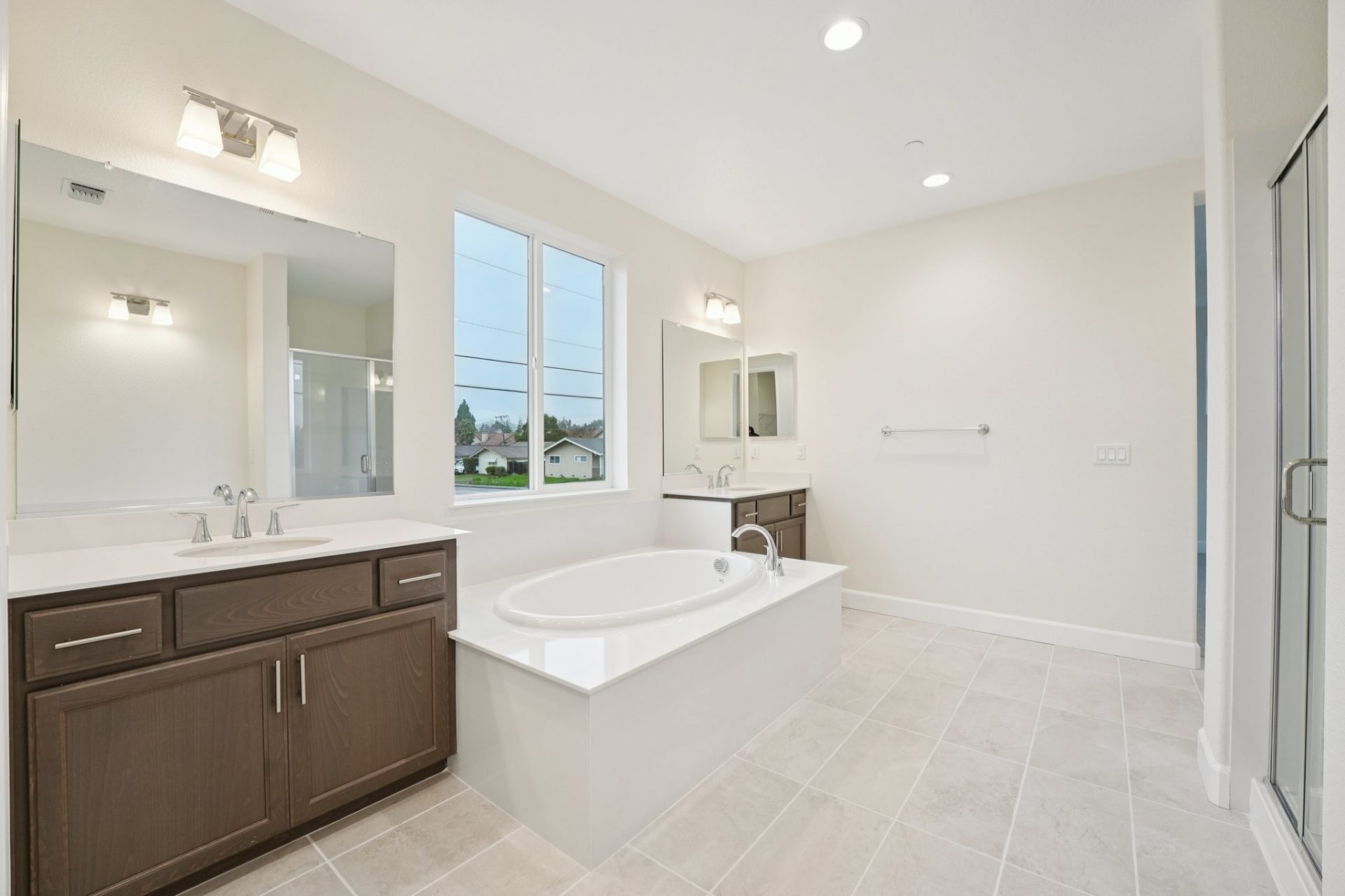 Bathroom with two separate vanities with sinks, and a soaking tub in the middle with a large window above it. A light colored tile on the floor, mirrors and lighting fixtures located above the vanities.