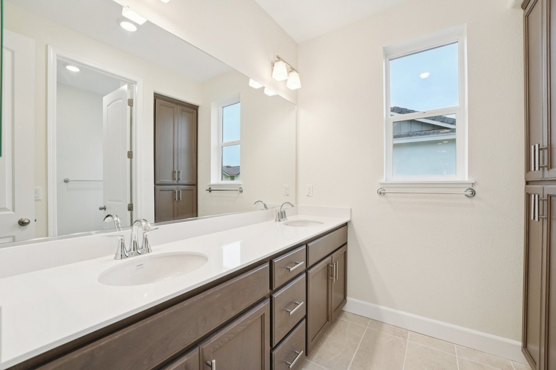 Bathroom with a dual sink vanity with brown stained cabinetry, nickel pulls, and nickel finish faucets. A light colored tile flooring, two-light fixtures and a window are seen.