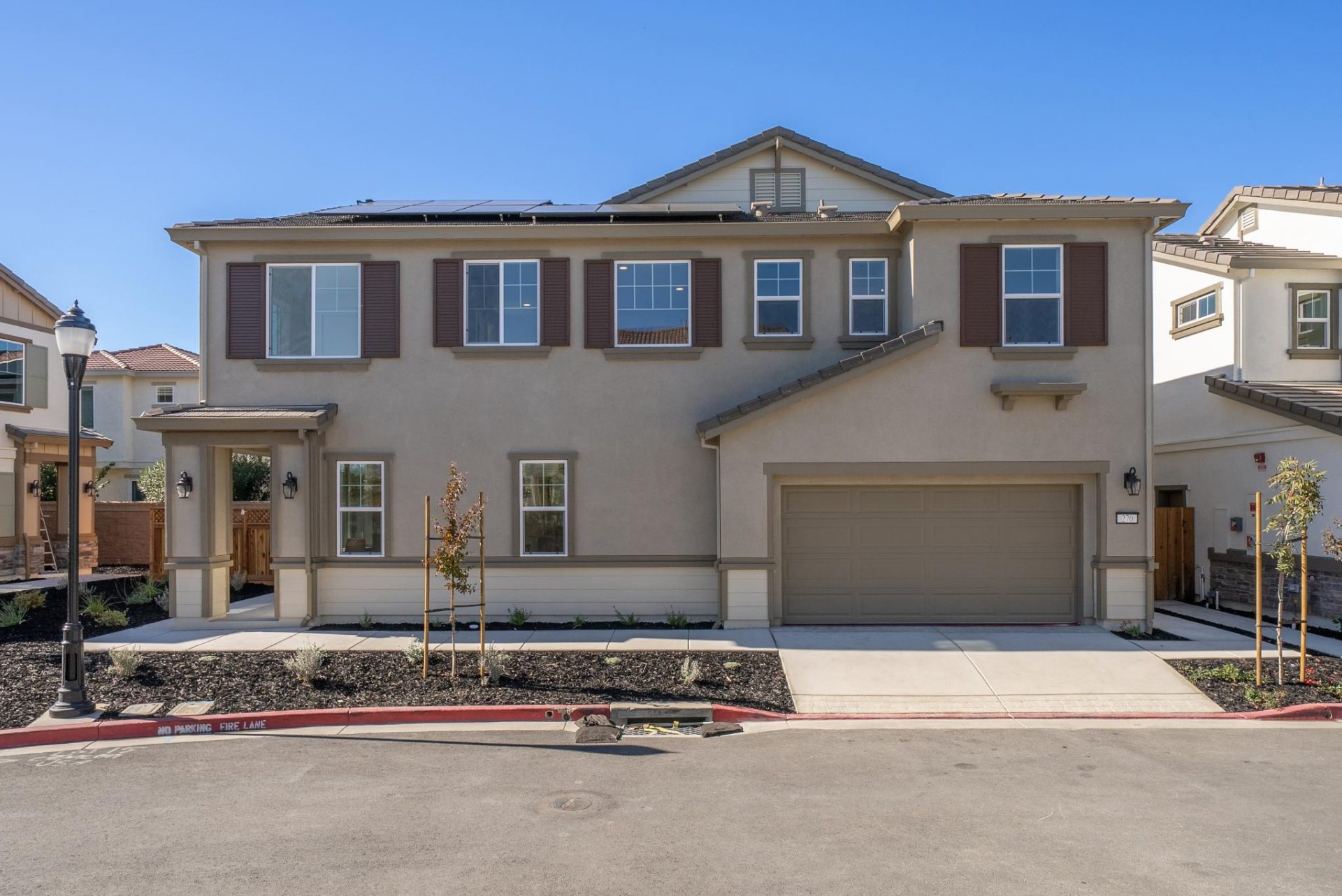 Exterior of two story home with neutral brown color scheme, window shutters, two car garage and bark mulch with trees and shrubs landscaping.