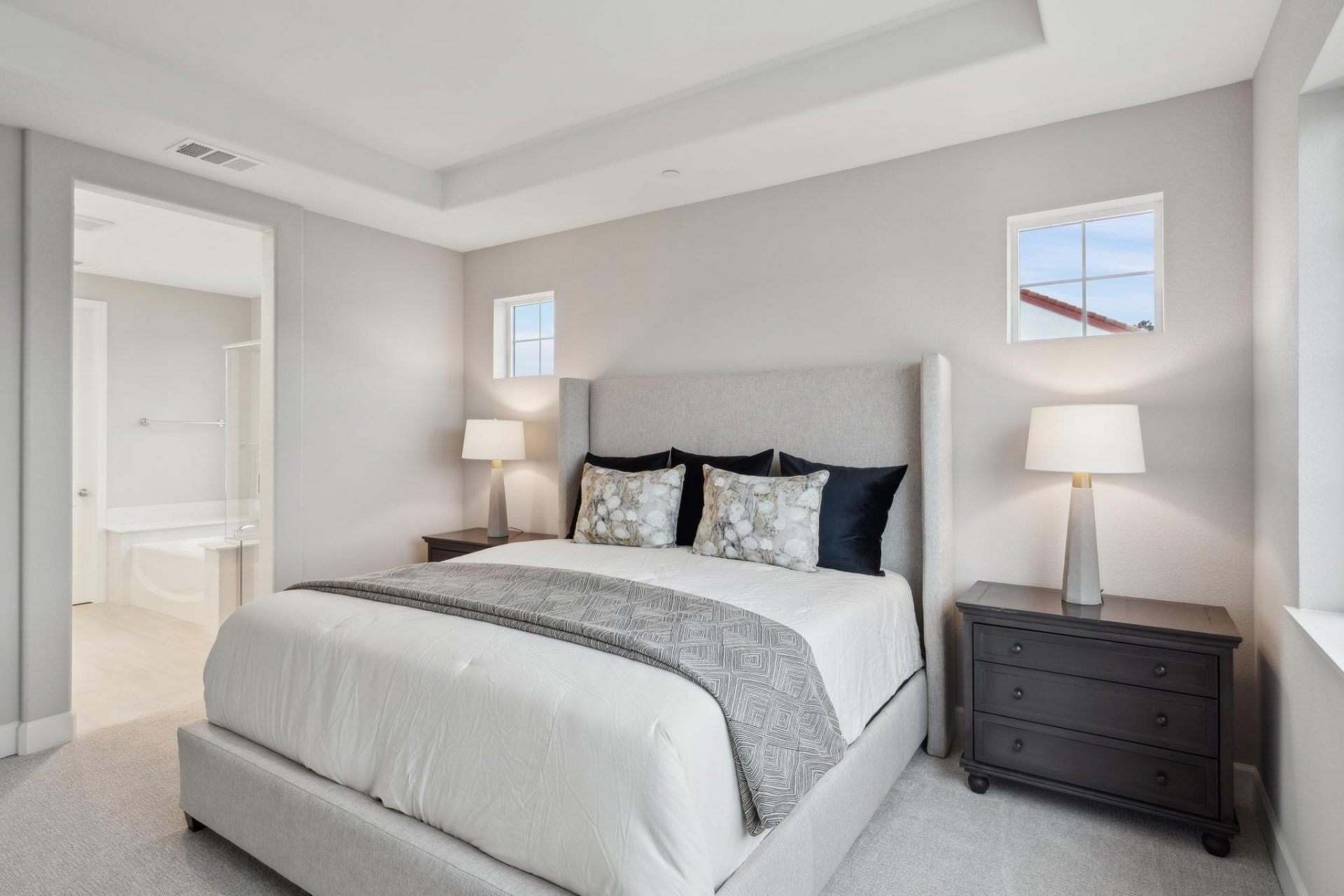 A staged bedroom with a headboard and rails two nightstands with lamps, beige, gray and black linens, medium gray walls and carpet and a bathroom seen through a doorway.