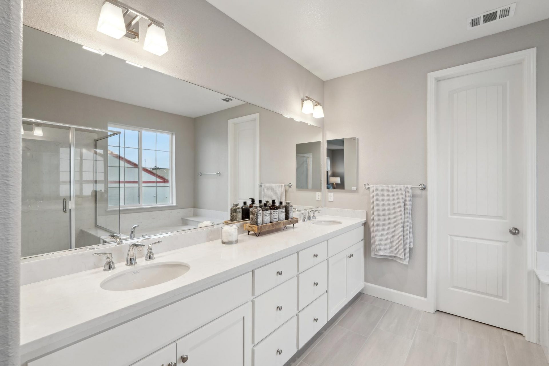 A bathroom with a long double sink vanity with white cabinets and nickel pull knobs, white countertops, gray walls, light gray floor tile and a long wall mirror with two light fixtures.