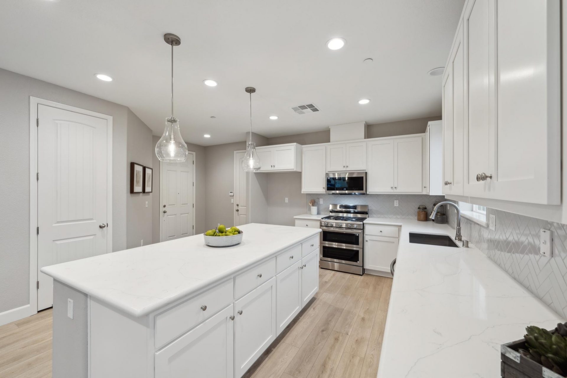 A kitchen area with an island with white cabinets and nickel pull knobs, white countertops, stainless steel appliances and glass pendant light fixtures over the island. Light wood flooring and gray walls with LED can lights in the ceiling.