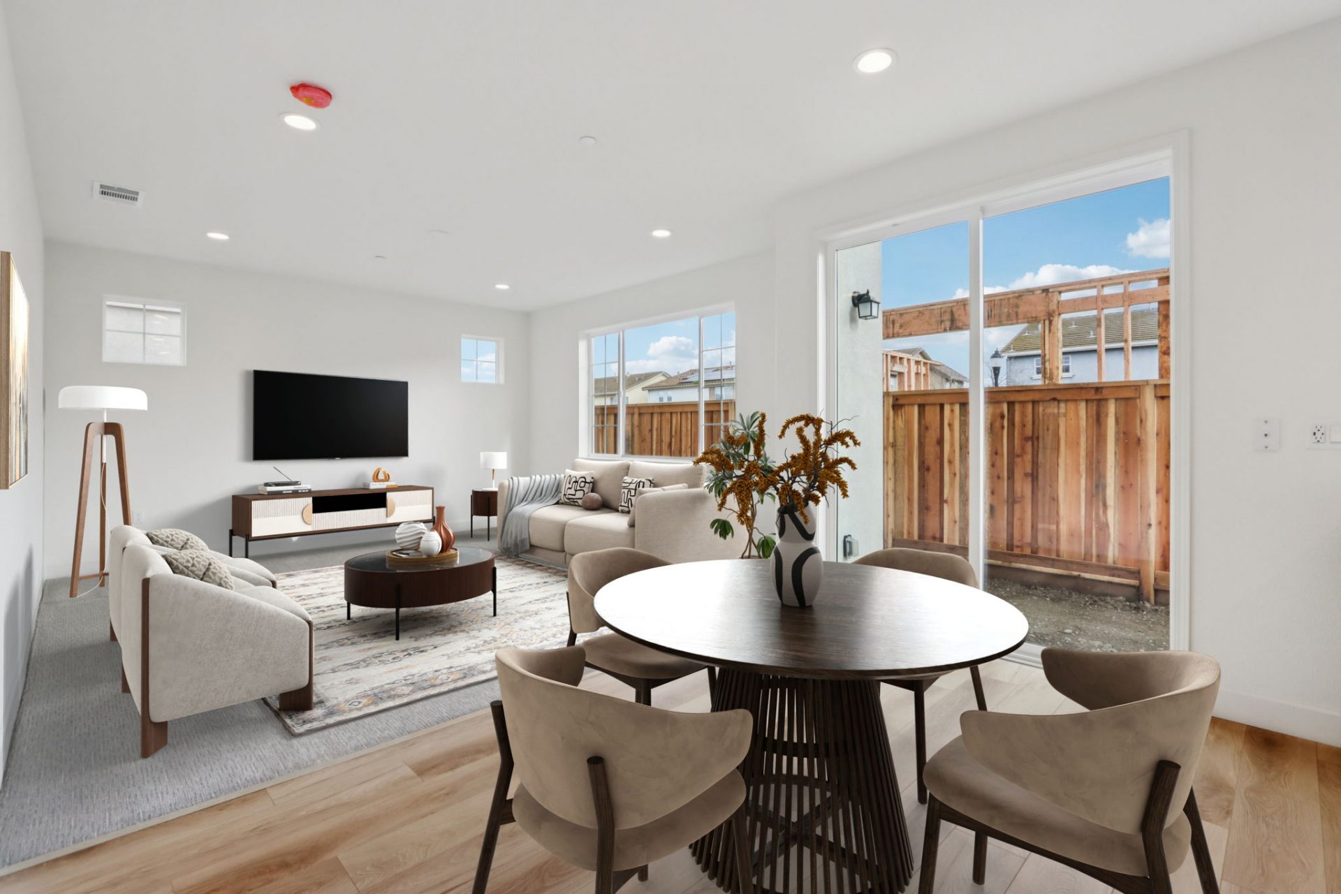 A virtually staged dining area with hardwood floors with a round table and chairs, and a carpeted living area with chairs, couches, a tv stand and a wall mounted tv.