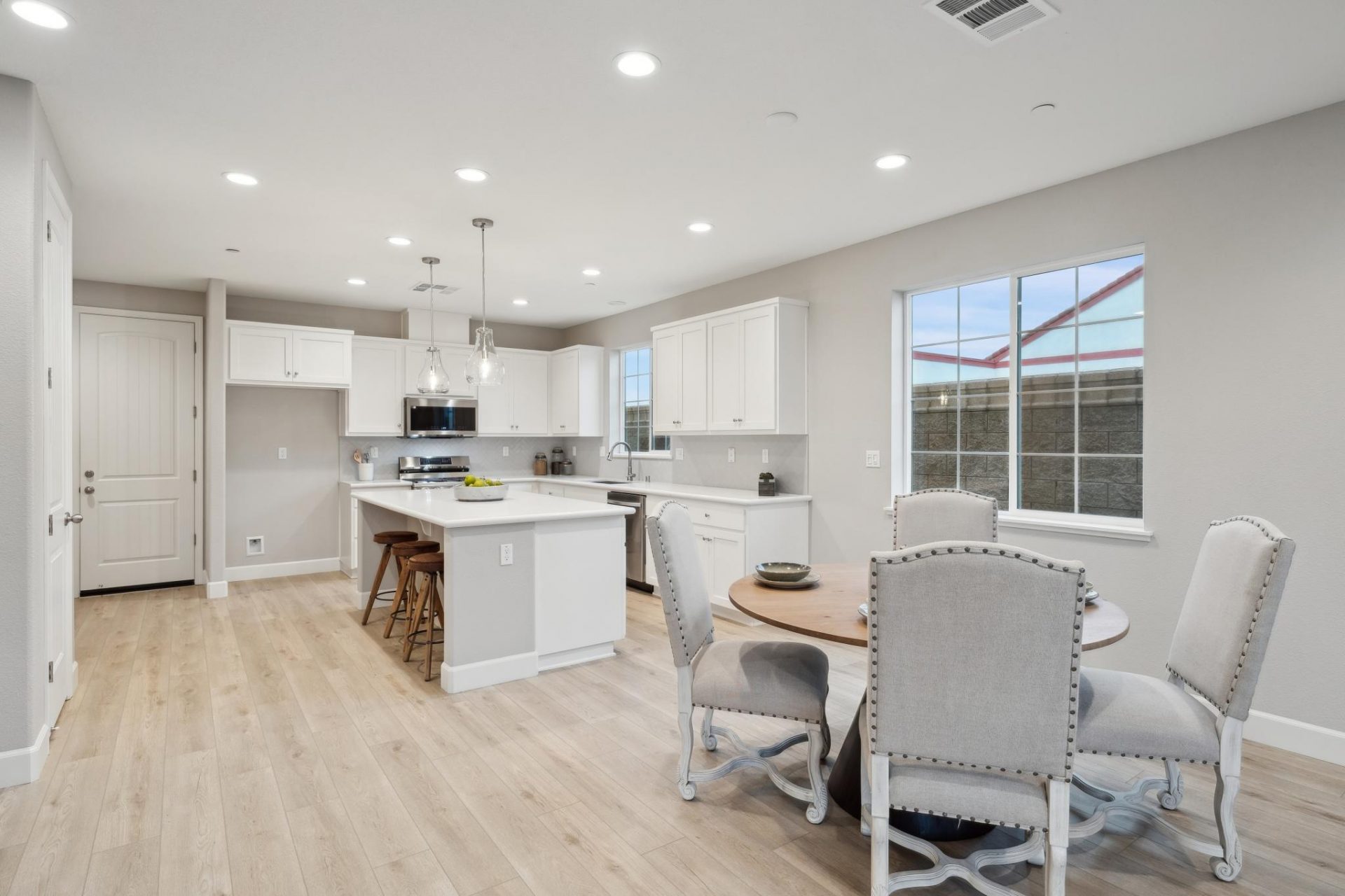 An open concept area with a dining table complete with four chairs, light wood flooring spanning across the space into the kitchen seen in the distance.
