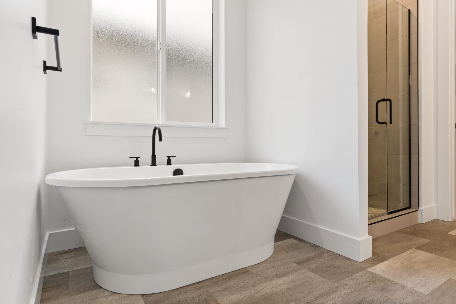 A free standing soaking tub with black faucet and black hardware with a glass enclosed shower space to the left. Medium brown floor tile and white walls.