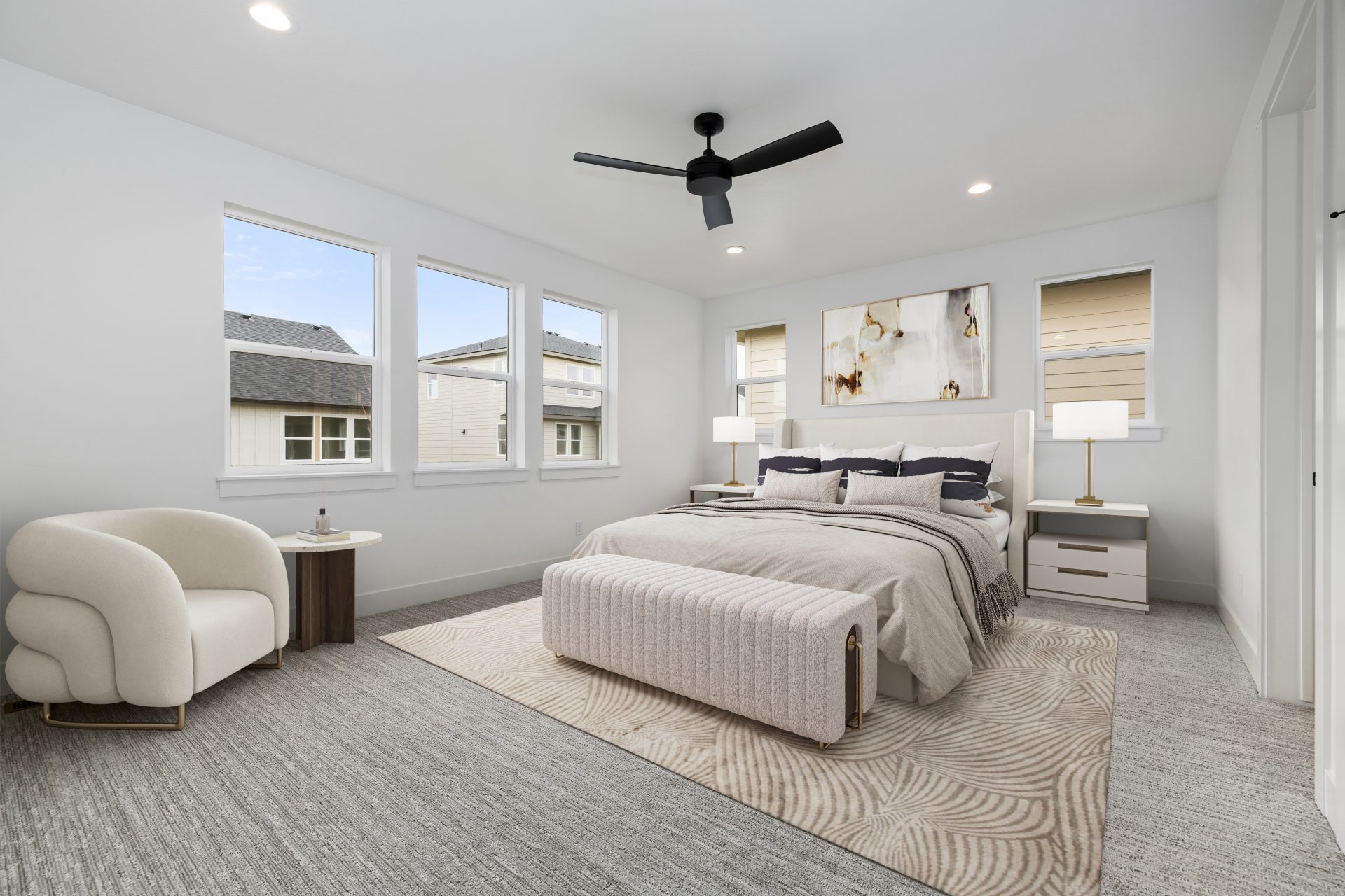 A virtually stage bedroom with neutral color headboard with rails and mixed neutral colored linens. At the edge of the bed a seating lounge and chair with small table, an area rug under the bed, two nightstands with lamps and a ceiling fan along with LED 