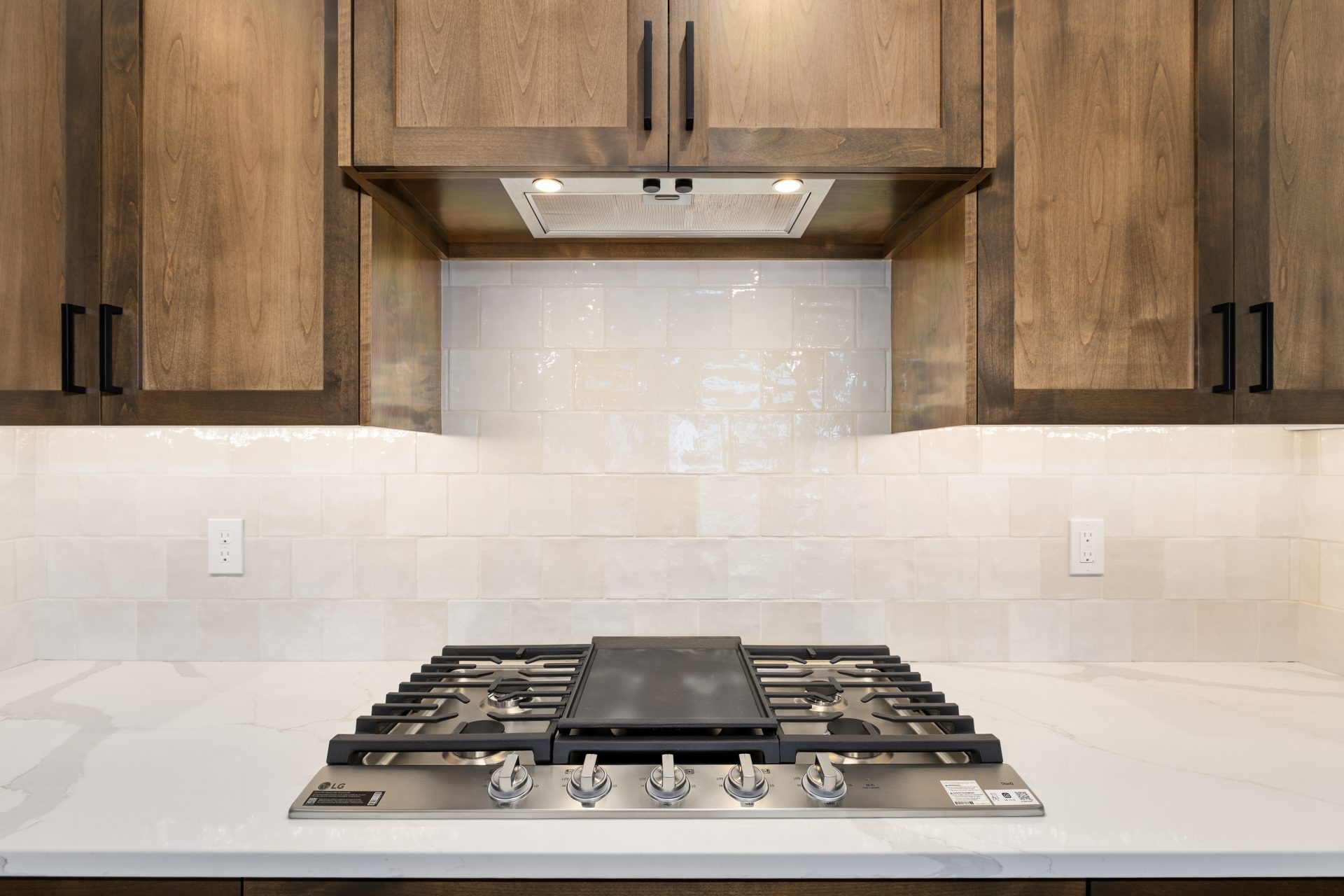 A stainless steel gas cooktop with white countertop to cabinet tile detail and brown stained cabinetry.