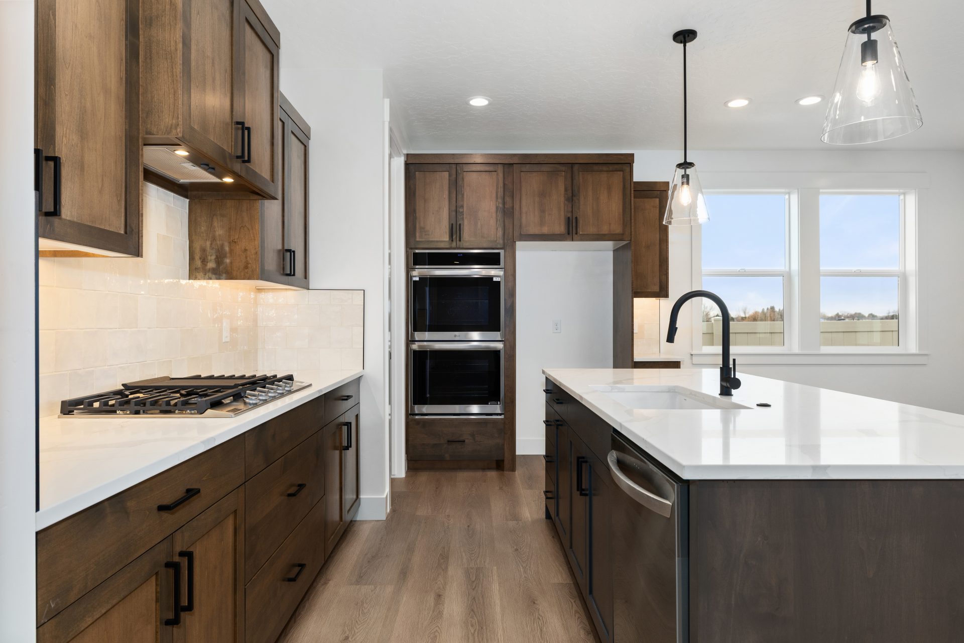 Kitchen view with kitchen island on the right, upper and lower cabinets with countertop and cooktop to the left. The cabinets are a dark brown stain and there are stainless steel appliances including double wall ovens, black finishes complete the space.