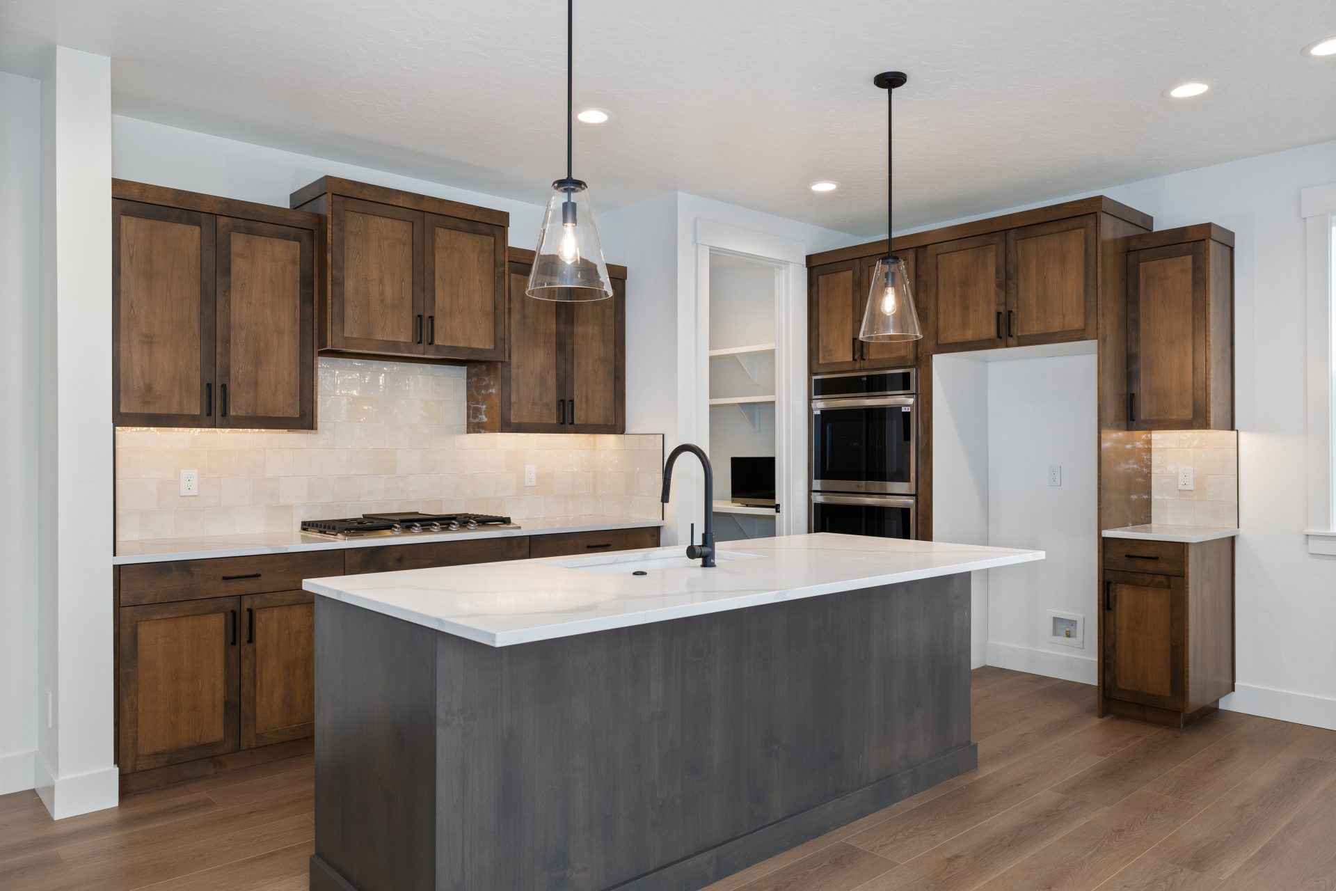 A kitchen with kitchen island, dark brown stained upper and lower cabinets, dual wall ovens, gas cooktop and white quartz countertops, a light color tiled backspace and glass pendent lights hanging over the kitchen island.