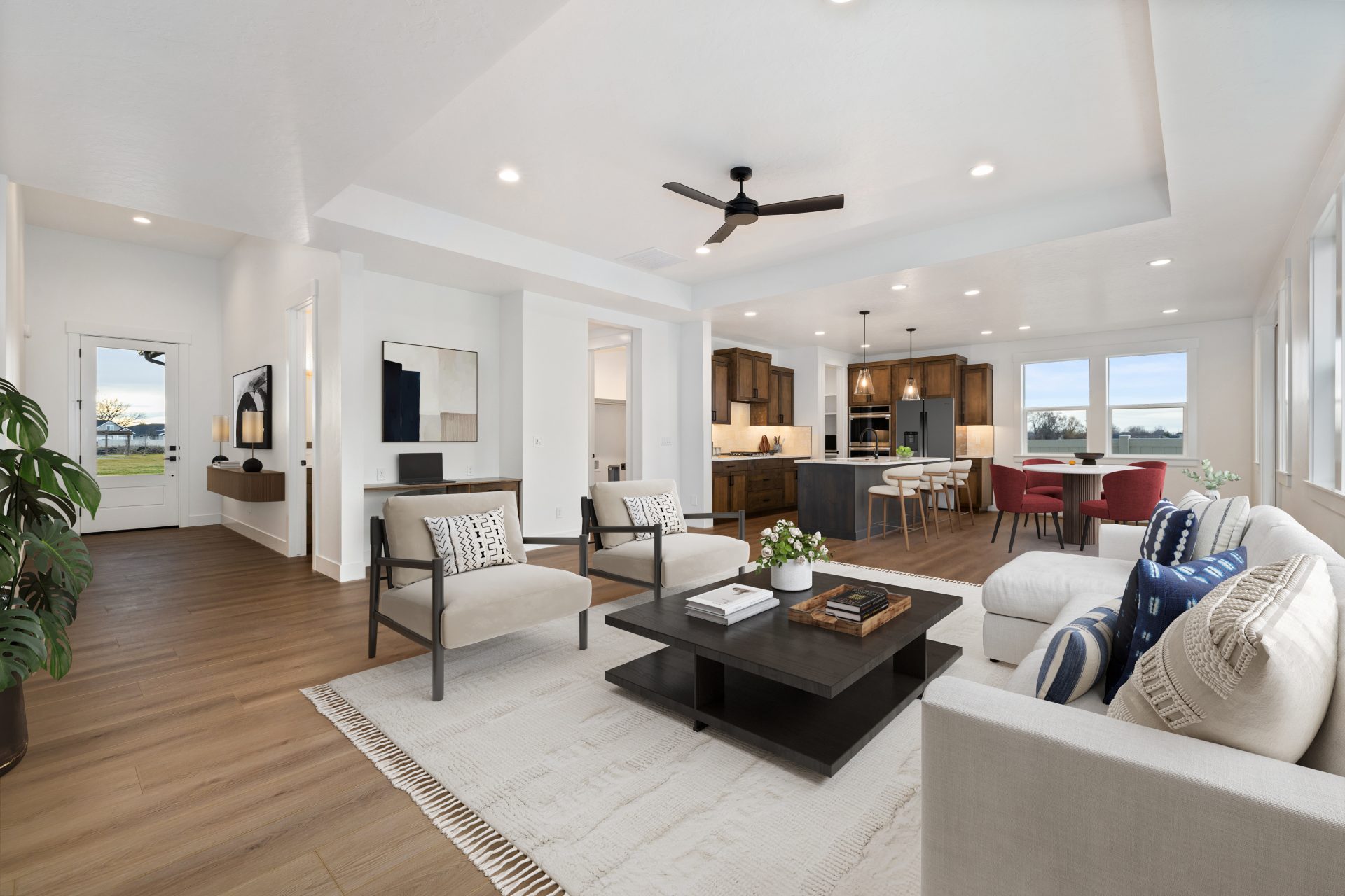 A virtually staged interior open concept floorplan with medium brown flooring, a couch with chairs and square coffee table, a ceiling fan and LED can lights in the ceiling. In the far sight a kitchen and a virtually staged dining area.