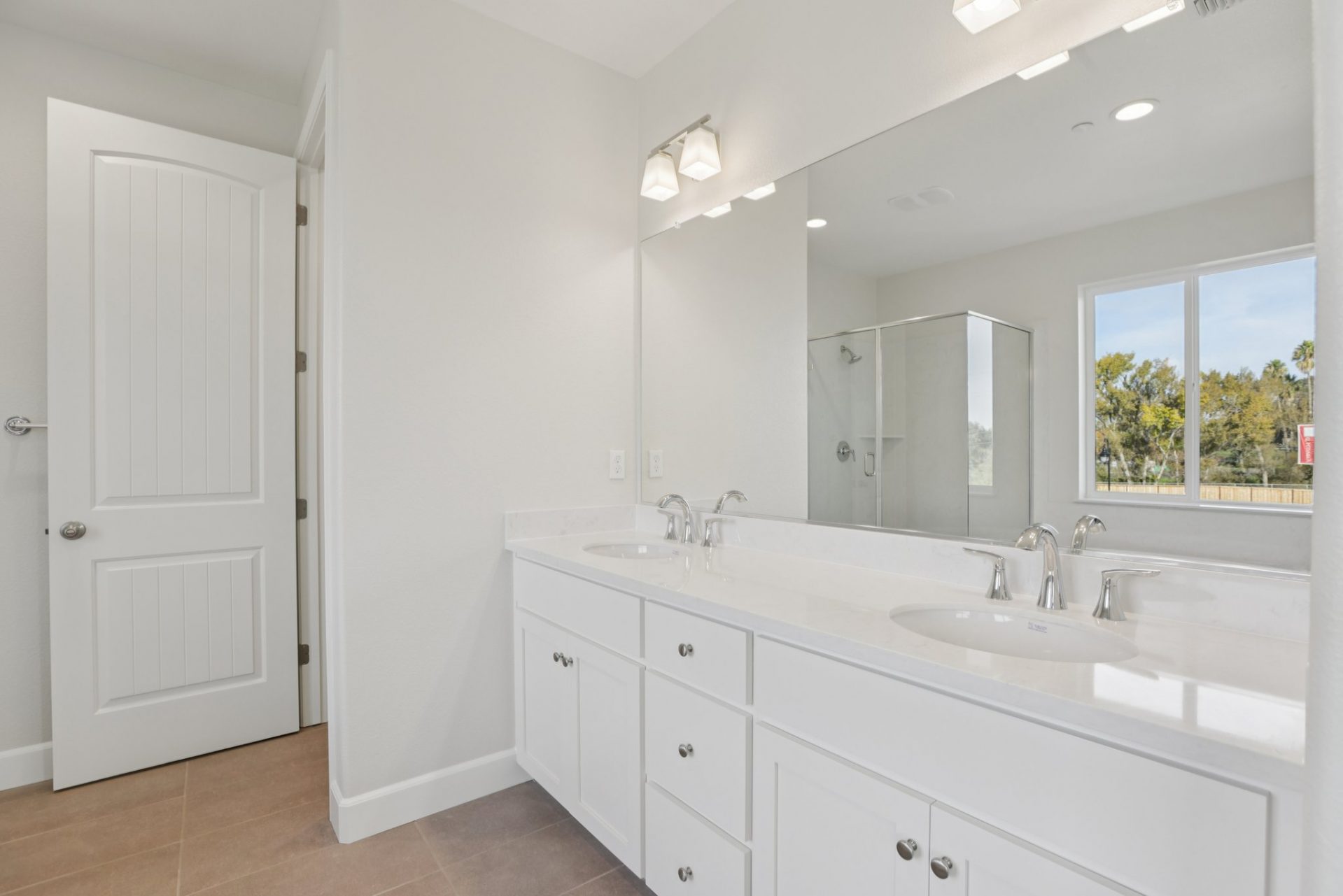 A two sink vanity with white cabinets, white counter top, chrome fixtures and a long wall mirror with two light fixtures above.