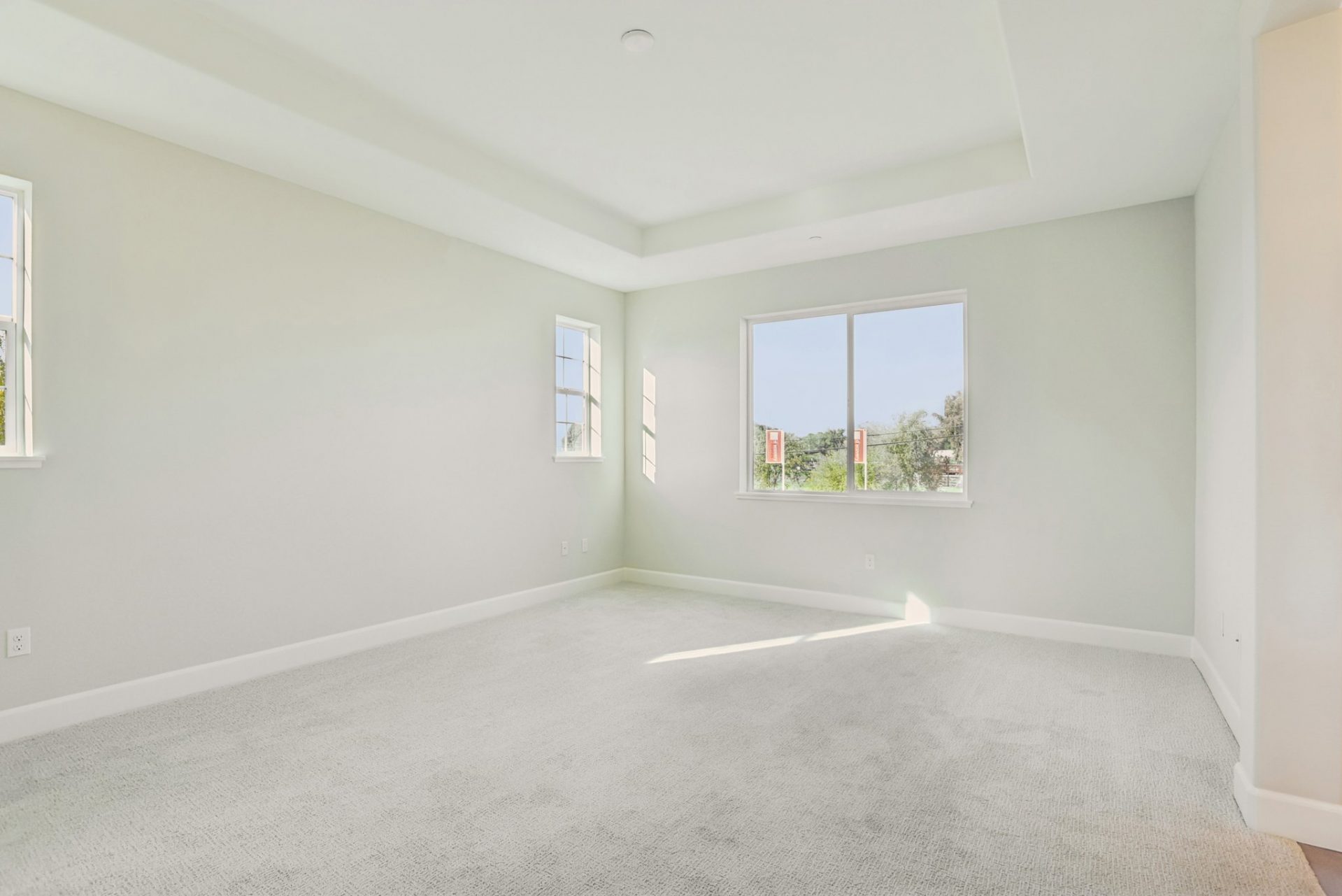 An un-staged room with light colored walls and carpet, three windows and an inset high ceiling.